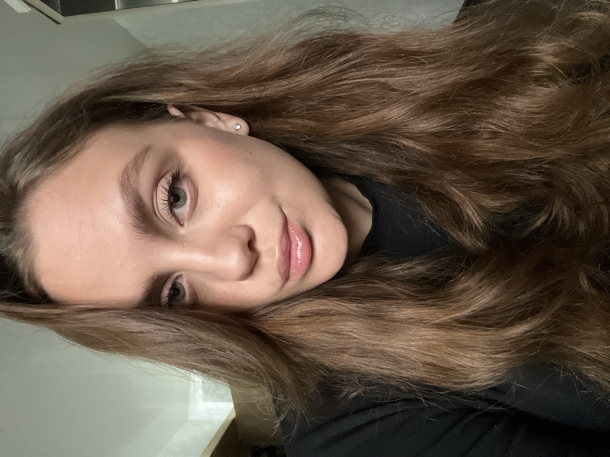 A young woman with long, light brown hair is seen in a close-up selfie. Her hair, styled in loose waves, fills much of the frame. She has fair skin, light green eyes, and full lips with a glossy finish. A small, round earring is visible in her left ear. She is wearing a black garment, visible at the lower right. The background is a plain wall and table. There is no information to conclude the picture was taken in the city of Chaikovsky, Russia.