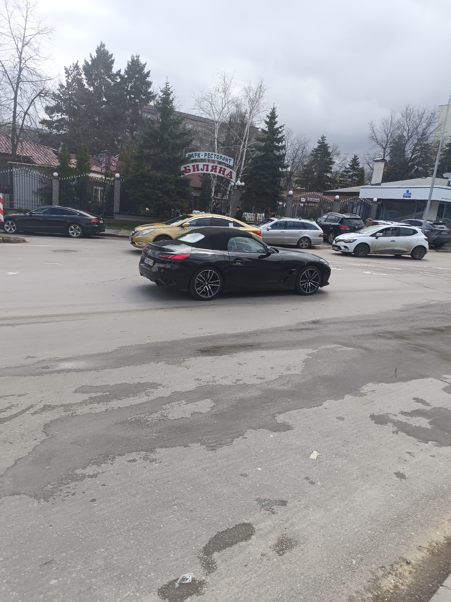 The image depicts a street scene in Sofia, Bulgaria. A black convertible BMW Z4 is in motion, moving left to right in the frame. Several other vehicles are parked on the side of the road, including a black sedan, a yellow taxi, a silver station wagon, a black SUV, and a white hatchback. The "Park-Restaurant Bilyana" signage is visible in the background. Across the street is a building with "OББ" (United Bulgarian Bank) signage. The street is paved and appears wet with patches of moisture. Trees and buildings line the background. The sky is overcast.
