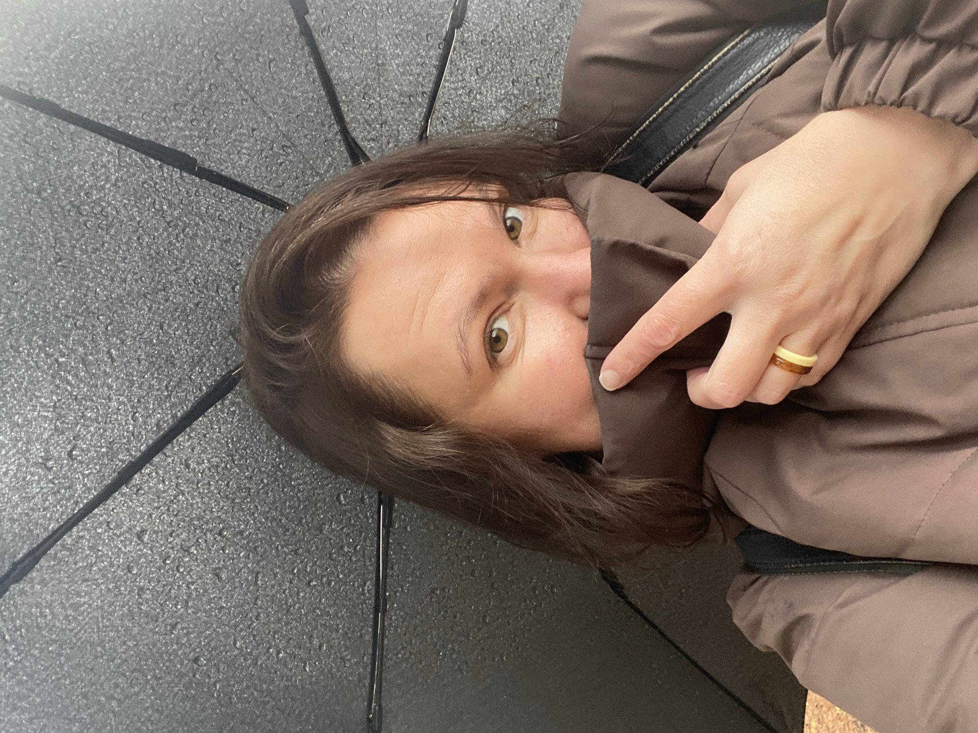 A person, likely a woman, looks directly at the camera from beneath a dark umbrella covered in numerous small water droplets, indicating rainy or wet conditions. Her head is tilted, and she has brown hair and prominent brown eyes that appear wide open. She is wearing a brown puffer jacket, with her right hand partially covering her face with the jacket's high collar, possibly for warmth or protection from the weather. A two-toned ring, featuring light yellow/beige and brown bands, is visible on a finger of her right hand. A black strap, potentially from a bag or part of the jacket, can be seen near her shoulder. The image is composed from a low angle, focusing on the person and the wet canopy of the umbrella overhead. The scene is set in Chaikovsky, Russia. There is no visible text in the image.