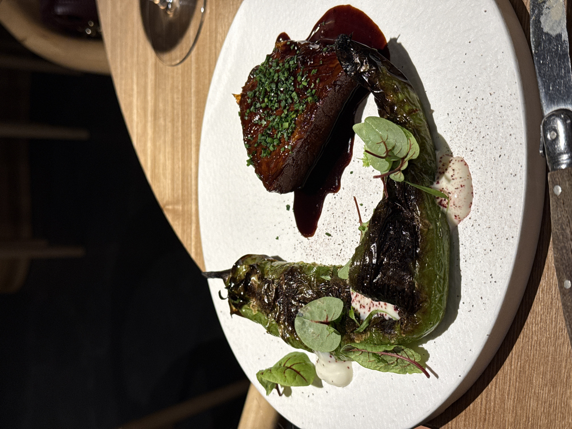 A culinary presentation is captured from an overhead angle on a white plate. The plate contains a dark-brown, rectangular piece of meat covered in a dark sauce and sprinkled with finely chopped green herbs. Adjacent to the meat is a char-grilled green pepper, curved in shape, adorned with small leafy greens and a white sauce speckled with red. A portion of a Laguiole knife rests on the plate's edge. The scene is set on a wooden surface, with shadows indicating directional lighting. The image may have been taken in a restaurant located in Krasnoyarsk, Russia, but this cannot be definitively determined from the visual content alone.