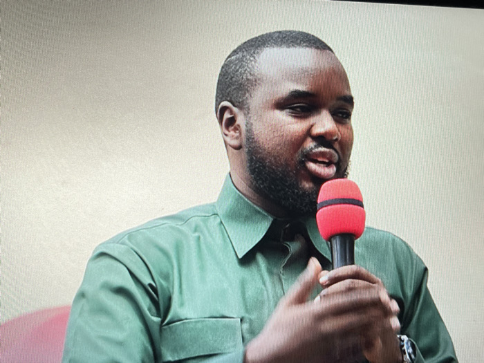 The image shows a Black man with a dark beard speaking into a microphone. He is wearing a green long-sleeved shirt and has a watch on his left wrist. The background appears to be a light-colored wall. The image was taken in Keshero, Democratic Republic of the Congo, at coordinates -1.648 latitude and 29.184 longitude.