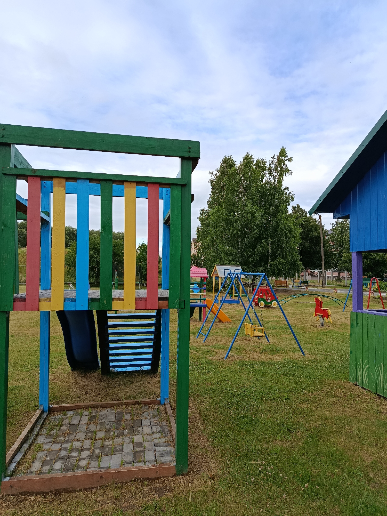 The image depicts a colorful children's playground on a cloudy day. The playground features various structures made of wood and metal, painted in bright primary colors. In the foreground, a wooden play structure with red, yellow, and blue vertical slats is prominent. Below this structure, a dark blue slide is visible, along with a section of horizontal blue and black slats.
To the right, a blue, red, and green playhouse with a pitched roof stands. In the center of the playground, a blue swing set with two yellow swings is set up. Behind it, a red and green climbing structure with a yellow slide is present. Further in the background, a red spring rider in the shape of a horse is visible, along with other play equipment and a bench. The ground is covered in green grass with some patches of dry grass and small white flowers. Trees and buildings can be seen in the distance. The sky is overcast with scattered clouds. The scene appears to be a public park or an outdoor play area for children, likely located in a residential or rural setting, given the presence of trees and distant buildings. There is no visible text in the image. The overall mood is peaceful and inviting, suggesting a place for recreation and fun.