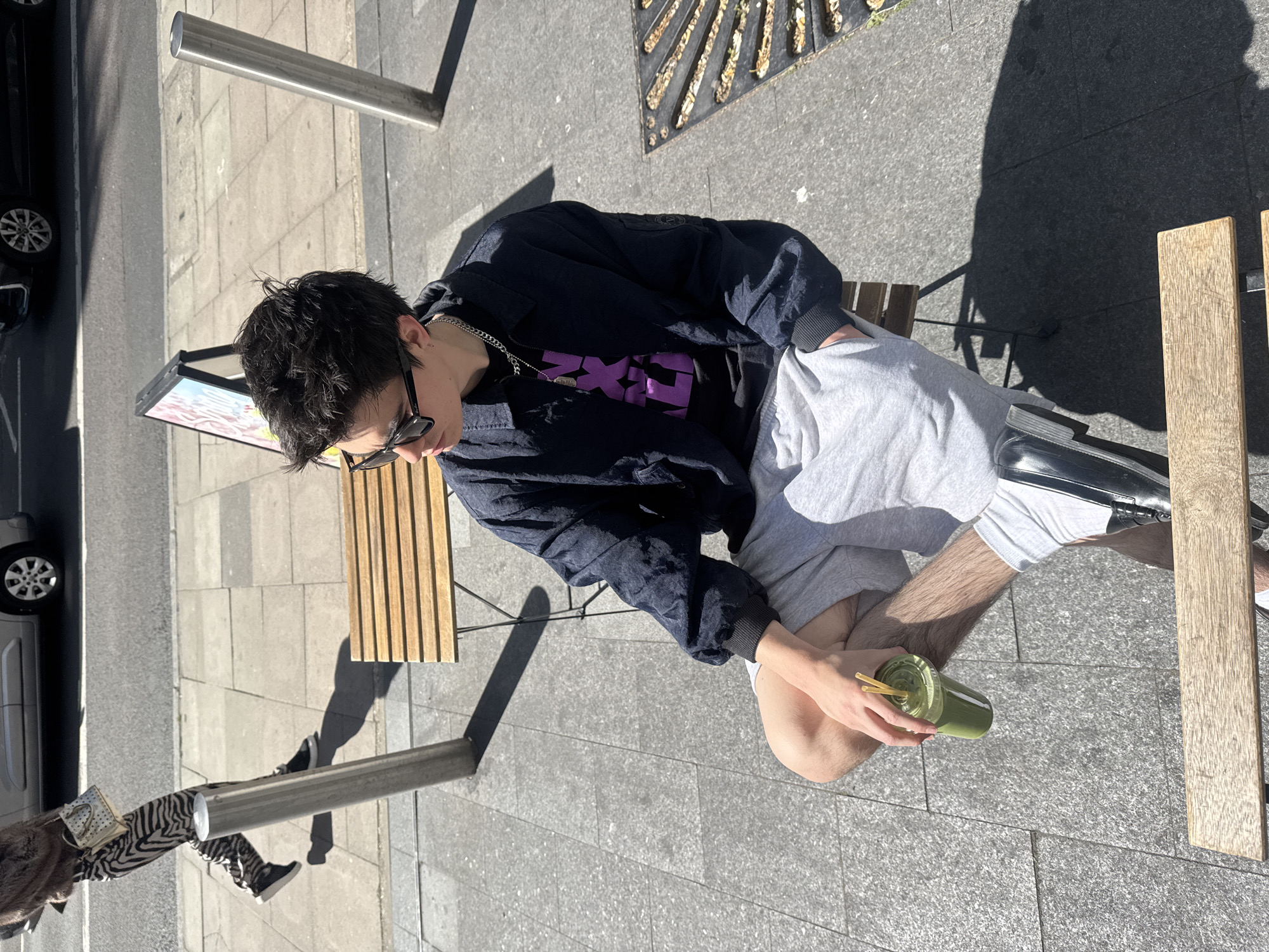 A young person is seated at an outdoor wooden table on a sunny day in London, United Kingdom. The individual has dark hair and is wearing black sunglasses, a dark blue or black jacket over a purple graphic t-shirt, and light grey shorts. They are holding a clear cup with a green beverage and two straws. The text visible on their t-shirt is "NXT LVL" in a stylized purple font.
The scene is an urban outdoor setting, possibly a cafe patio or sidewalk, with grey paved ground and strong shadows cast by the bright sunlight, indicating clear weather during the day. In the background, a small advertising stand is visible. Further back, part of a black car can be seen on the street. Another person, partially visible, walks past in the background, wearing zebra-print pants and holding a light-colored handbag. Metal poles are also present in the environment. The person seated appears relaxed, enjoying their drink in the bright outdoor setting.