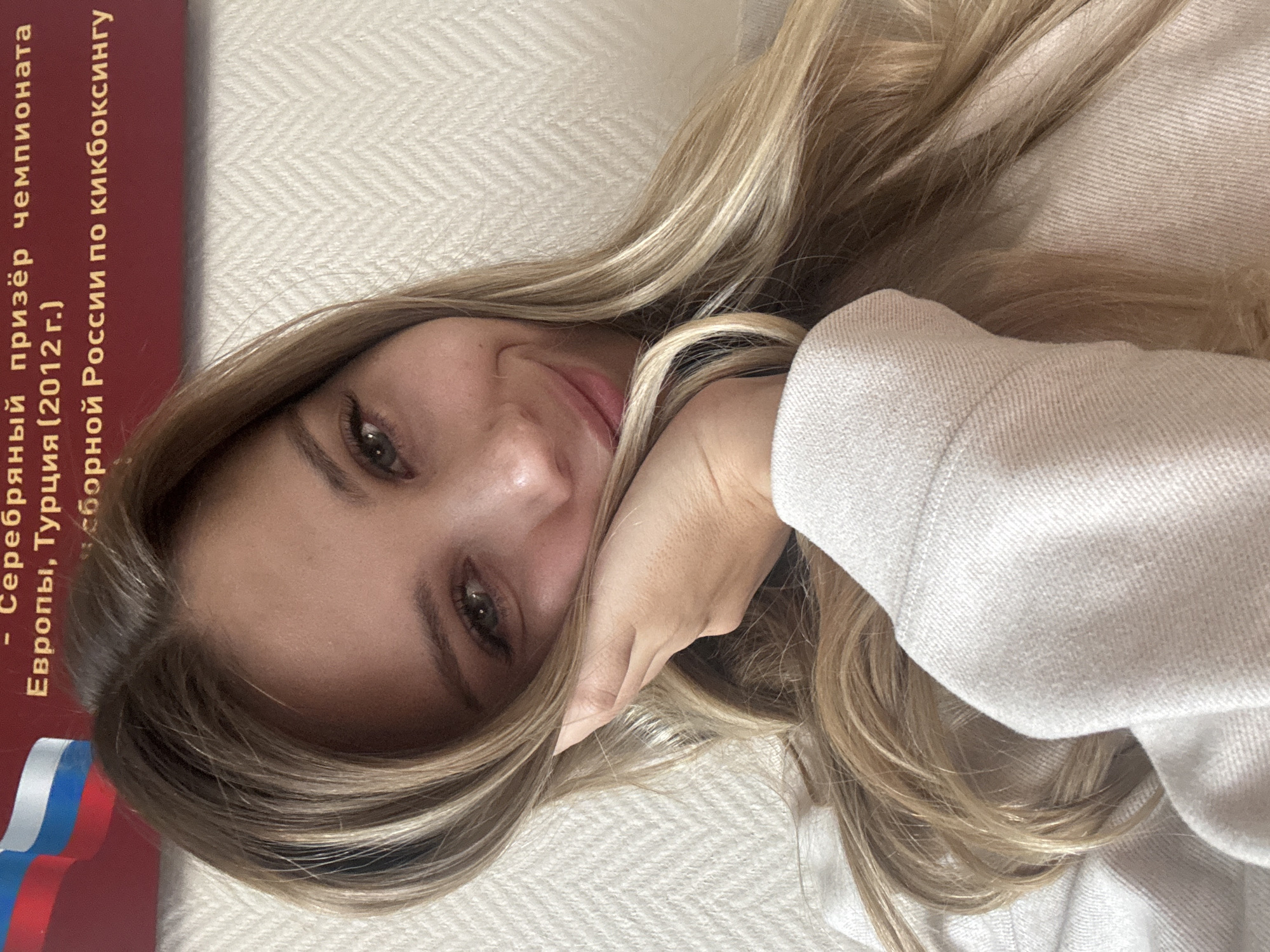The image shows a close-up of a young woman with blonde hair, resting her head on her hand and smiling. She is wearing a light-colored sweatshirt. In the background, there is a textured wall and a portion of a red banner with Russian text and the Russian flag. The text on the banner appears to relate to a championship and mentions "Europe, Turkey (2012)" and "Russian kickboxing team". The presence of the Russian flag and the text referencing a championship and a specific location (Europe, Turkey) suggests that the image might have been taken in Russia, potentially in a city like Chaikovsky, where such sporting events or recognitions might be displayed. However, the image itself does not contain direct visual cues to confirm the exact location within Chaikovsky.