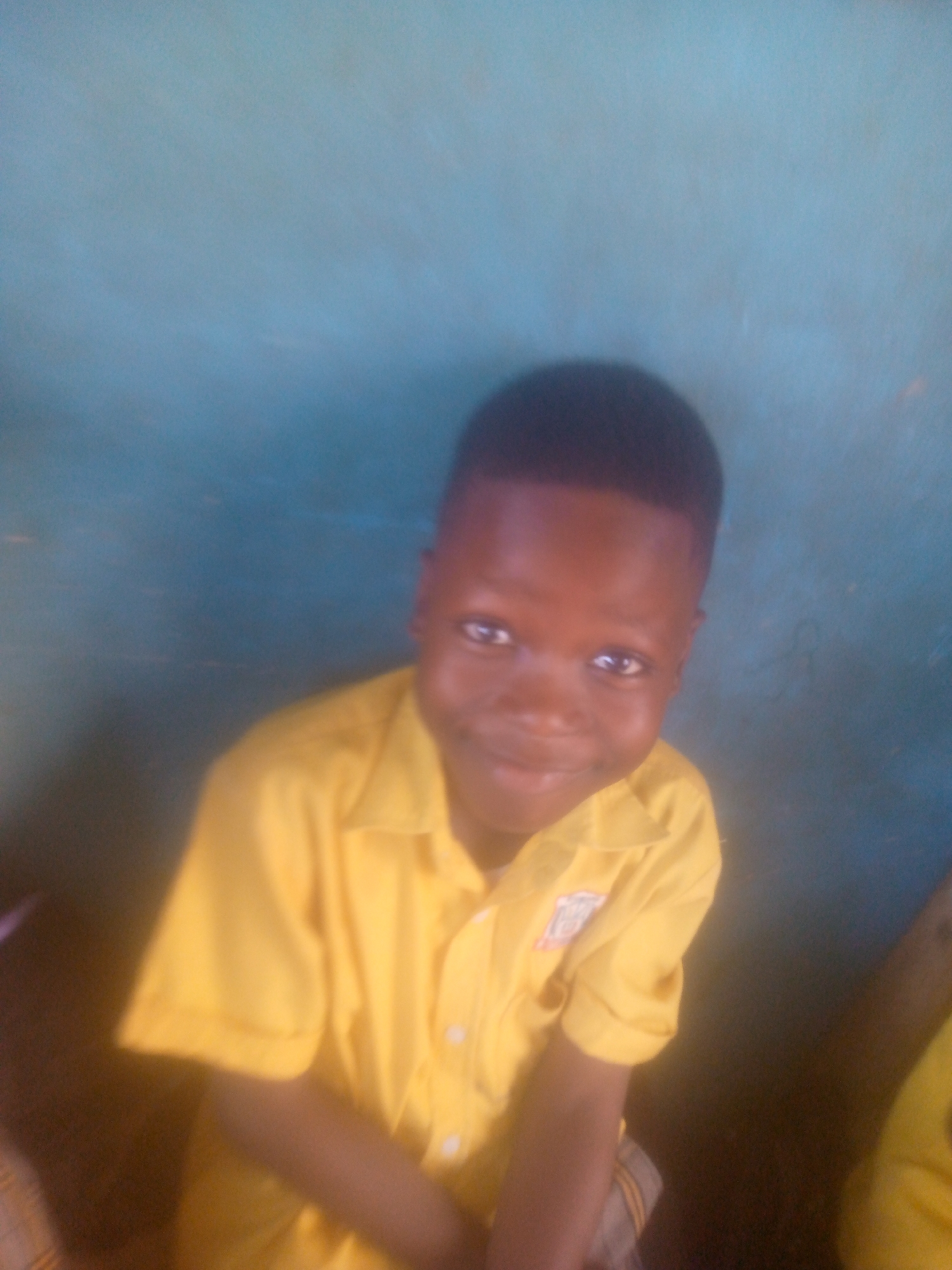 A young boy with dark skin and short, dark hair is smiling at the camera. He is wearing a yellow, short-sleeved button-down shirt. The background is a mottled blue color. The location is in the city of Unknown, Unknown.