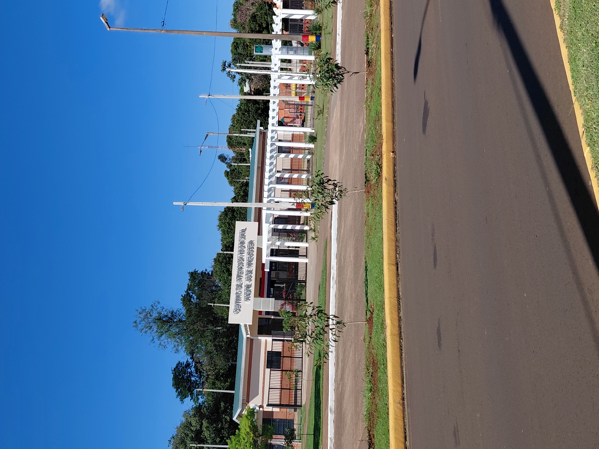 The image shows the exterior of the Centro de Atención Municipal Andre José Kresser in Encarnacion, Paraguay. The building has a light-colored facade with some brick accents and a green roof. A prominent sign above the entrance reads "CENTRO DE ATENCION MUNICIPAL ANDRE JOSE KRESSER". In front of the building, there is a white pergola structure supported by columns, and behind it, a fence encloses a courtyard with some playground equipment visible. The grounds are landscaped with grass, trees, and a yellow curb along the adjacent paved road. Several streetlights are visible in the background against a clear blue sky. There are no people visible in the image.