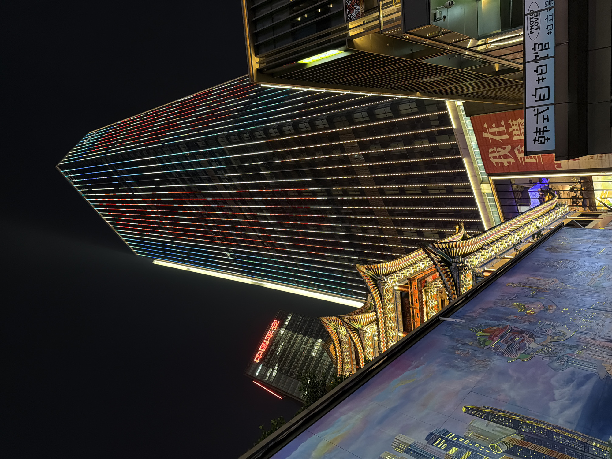 The image shows an upward view of a city scene at night in Yuzhong District, China. A tall, modern skyscraper with illuminated horizontal lines of red, white, and blue lights dominates the left side of the frame. To the right and slightly below the skyscraper, there is a traditional Chinese-style building or structure with ornate, golden-lit details and a curved roof. Below this structure, there is an image, likely a mural or display, depicting a futuristic cityscape. In the upper right, signs with Chinese characters, "PHOTO LOVE" and "韓式自拍馆 帕拉丁店", are visible on the side of a building. Further down a banner with more Chinese characters reading “我在麒麟” can be seen. In the bottom left corner, part of another skyscraper is visible, illuminated with red Chinese characters at the top. The sky is dark, suggesting it is nighttime.