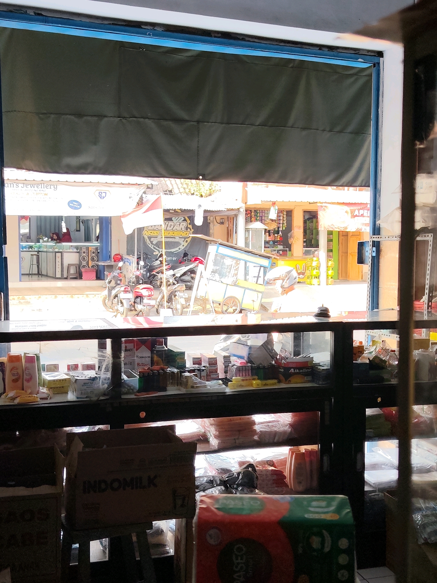 This image depicts the interior of a shop in Dampit, Indonesia, viewed through an open, dark green canvas shutter. The foreground is dominated by shelves filled with various products, including packaged goods, bottles of liquid, and boxes. One prominent box in the lower center is labeled "INDOMILK." Behind the shelves, through the open shutter, a street scene is visible. This includes parked motorcycles, a food cart, and storefronts with signage, suggesting a commercial area. The lighting indicates it is daytime. There are no discernible interactions between people or specific actions being performed in this static scene.