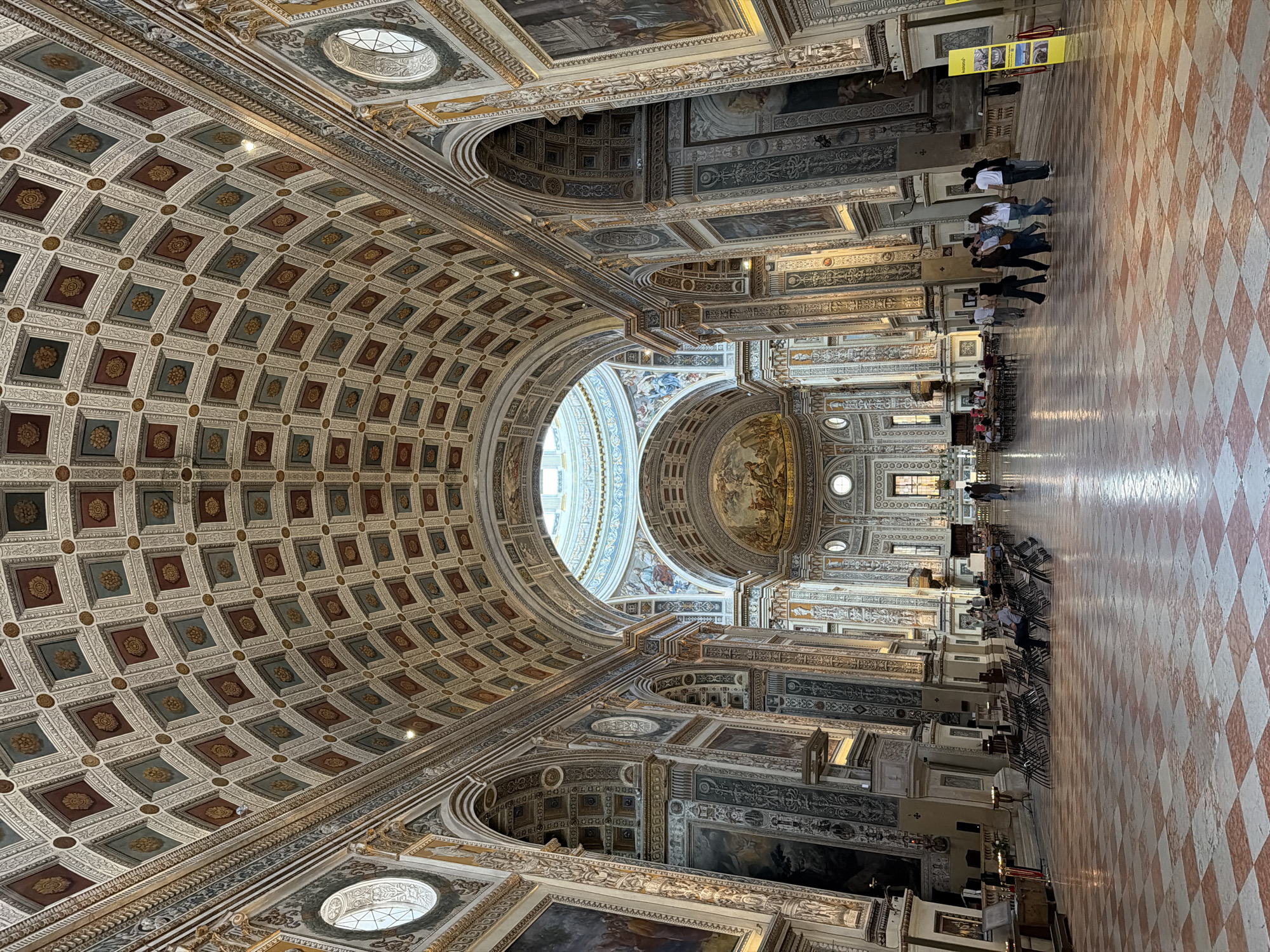 The image captures the interior of the Basilica di Sant'Andrea in Mantua, Italy, showcasing its ornate architecture. The ceiling is heavily decorated with a coffered design, and a large dome sits at the center. The walls are adorned with paintings and architectural details, including arches and columns. Several people are walking or standing within the basilica. The floor is covered in a red and white checkered pattern. Sunlight filters into the basilica through various openings.