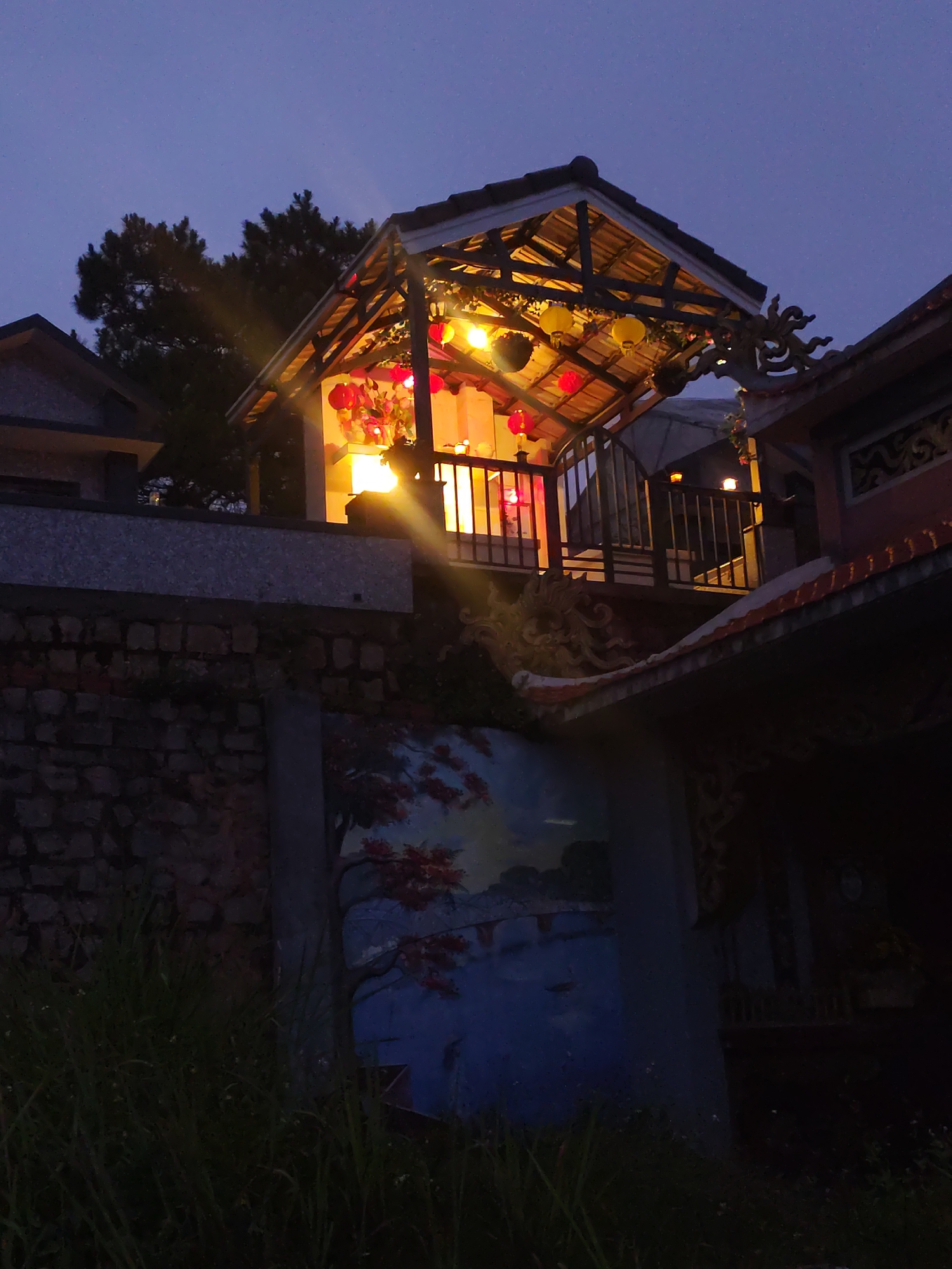 A low-angle, outdoor shot depicts a pagoda-like structure illuminated with hanging yellow and red lanterns. The structure has a tiled roof and decorative dragon-like carvings. The structure sits on a stone foundation with a mural of a blue landscape featuring a tree with red leaves. Tall grass grows in the foreground. Adjacent to the pagoda-like structure is another building with a red-tiled roof. A tree is on the left. The sky is a dark blue. The location is assumed to be in Lạc Dương, Vietnam.