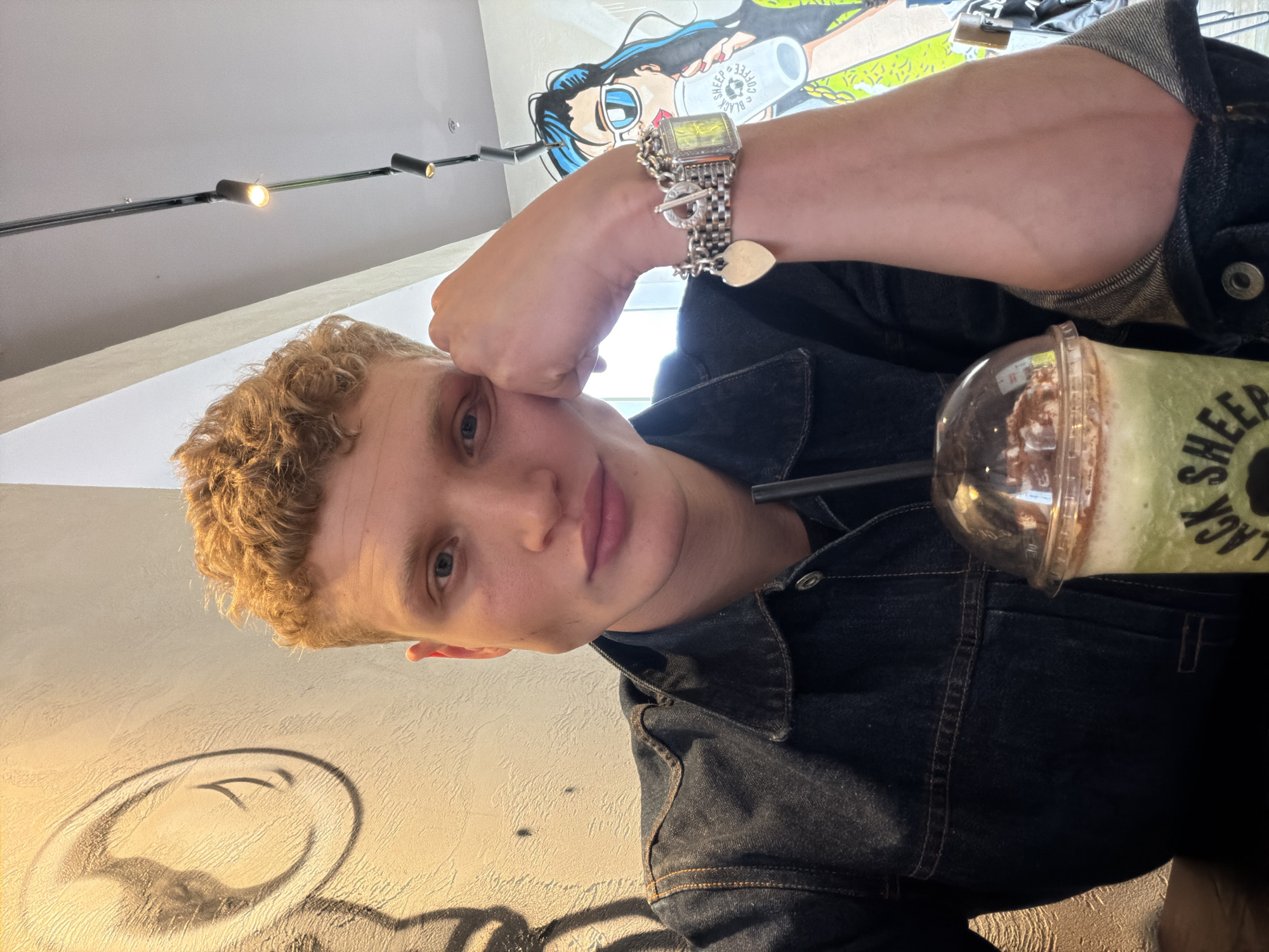 A young white male with blond, curly hair is seated at a table, leaning his head on his hand, and looking into the camera. He is wearing a dark denim jacket over a black t-shirt. On his left wrist, he wears a chunky silver bracelet with charms, including a heart and a lantern. A plastic cup with a green and brown beverage and a clear plastic lid is in the foreground, to the right of the subject. The background features a light-colored wall with a black mural to the left and a colorful mural of a person holding a coffee cup to the right. Overhead, a track lighting system with multiple spotlights is visible against a white ceiling. The scene appears to be an indoor cafe or restaurant, possibly in London, United Kingdom, given the style of the decor and the general ambiance.