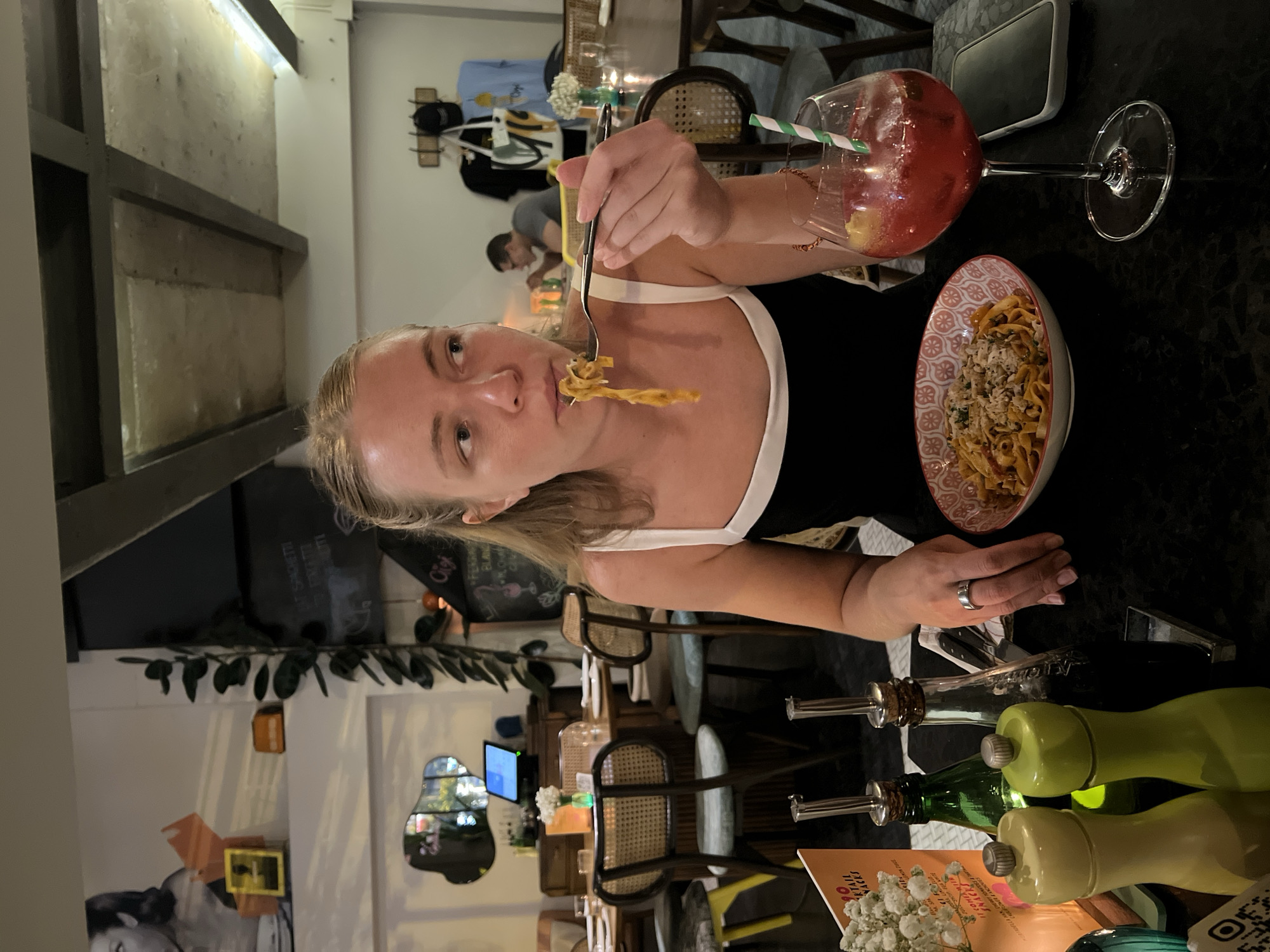 A fair-skinned woman with blonde hair is eating pasta in a restaurant. She wears a black top with white straps and holds a fork with pasta on it near her mouth. A bowl of pasta is to her right, along with a large stemmed glass containing a red drink with a straw. A black object resembling a phone or wallet lies beside the glass. The table is black and has a scattering of bottles and a menu. In the background, other tables, chairs, and patrons are visible, suggesting a casual dining atmosphere. The location is the city of Bangkok, Thailand.