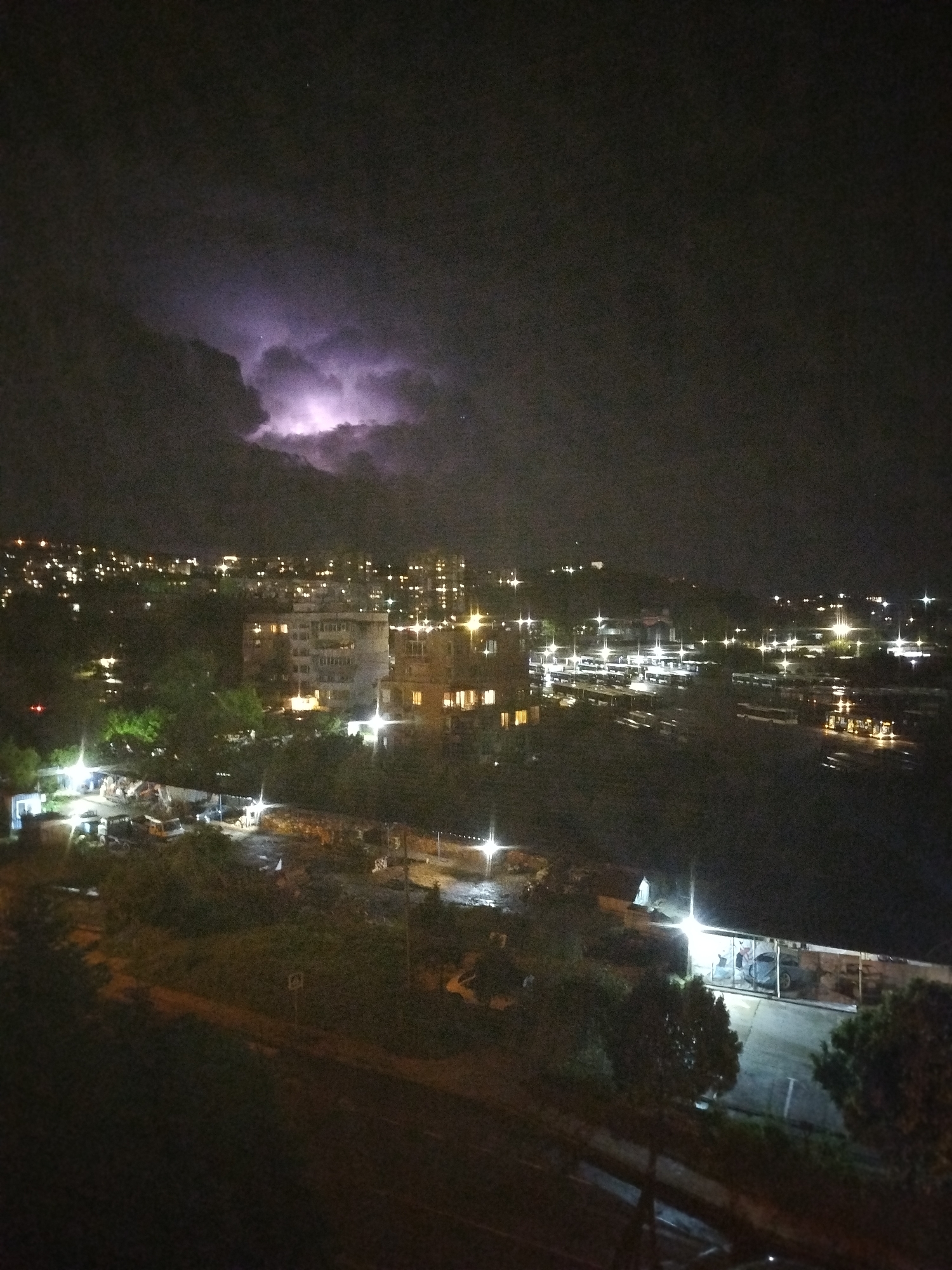 The image shows a night view of Varna, Bulgaria, with lightning in the sky. The city lights are visible in the distance, and there are several buildings and trees in the foreground. A bus depot with several buses is prominently visible in the middle ground. The environment is dark, with a thunderstorm occurring.