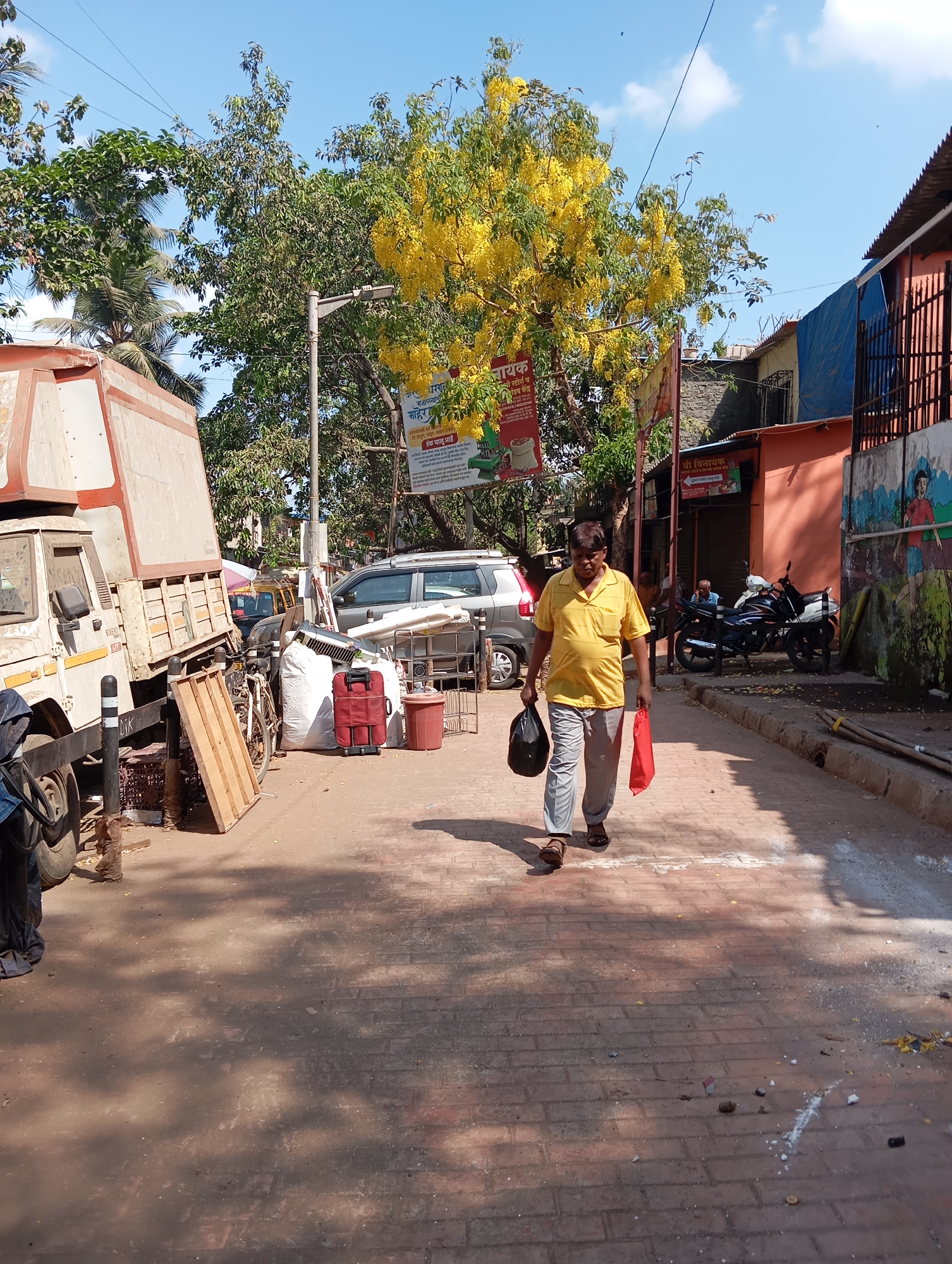 A man in a yellow shirt and gray pants walks on a red brick street in Mumbai, India. He carries a black bag in his left hand and a red bag in his right. The street is lined with buildings, vehicles, and various objects. A large truck is parked on the left side of the street, partially blocking the view. A tree with bright yellow flowers stands to the right, partially obscuring a sign. Two motorcycles are parked in front of an orange building. A mural of people playing is on a wall behind the building. The sky is bright blue with a few scattered clouds.