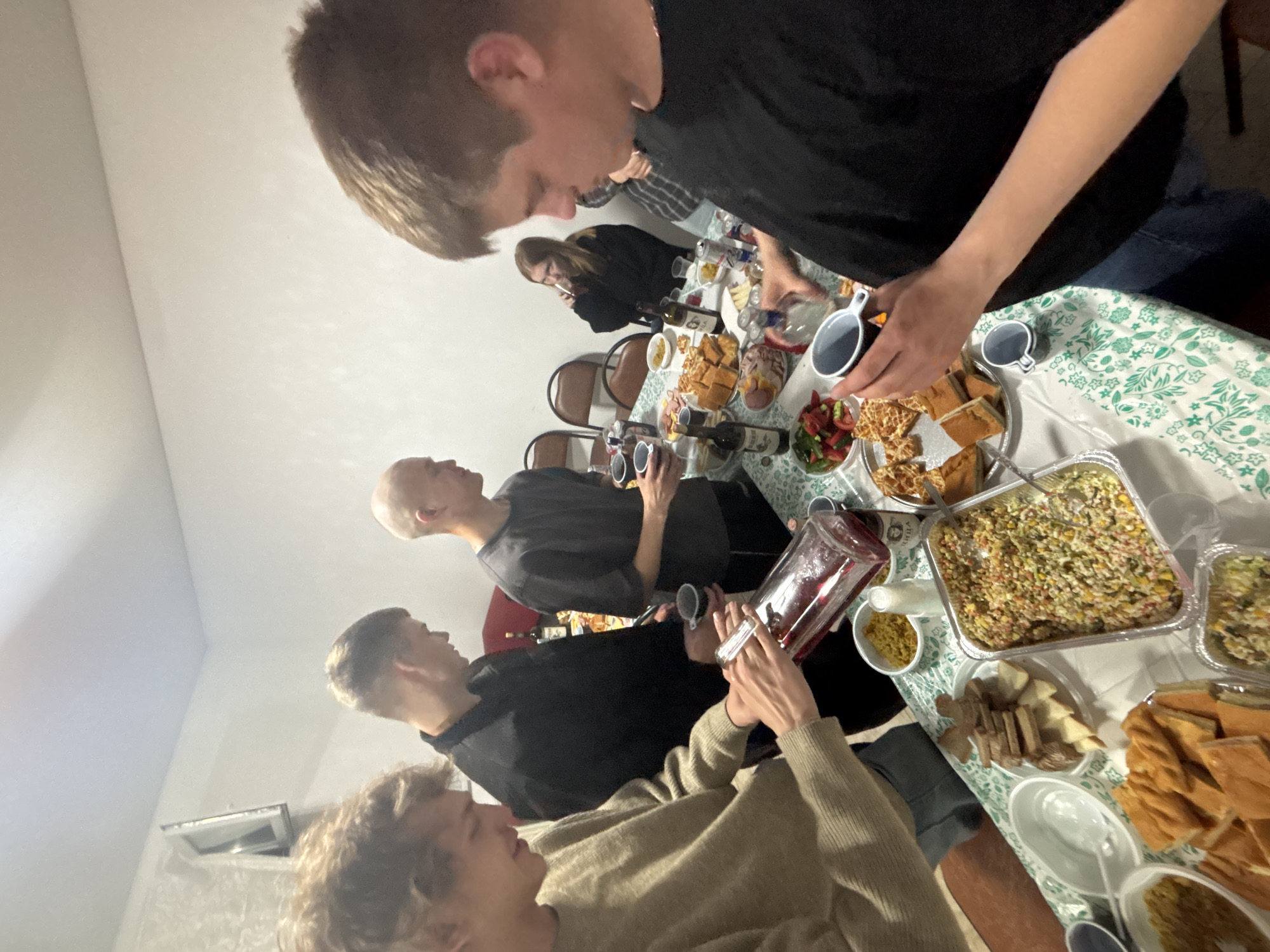 The image depicts a group of young adults gathered around a table laden with food and drinks, suggesting a social gathering or party. The setting appears to be an indoor, casual environment, possibly a home.
**People:**
There are at least five individuals visible, mostly young men.
* A young man on the far left, with light brown curly hair, is wearing a light brown sweater and pouring a dark red liquid from a glass pitcher into a cup.
* Next to him, a young man in a black long-sleeved shirt is looking down at the table, holding a cup.
* A bald young man in a grey t-shirt is also looking at the table and holding a cup.
* In the background, a young woman with long blonde hair is seated and holding a cup to her lips, seemingly drinking.
* On the right, a young man with short brown hair, wearing a black t-shirt, is holding a cup and a bottle, appearing to pour a clear liquid.
**Objects and Actions:**
* The table is covered with a green and white patterned tablecloth.
* Various food items are spread across the table, including what appears to be a large salad in a foil container, sliced bread, cheese, and other appetizers.
* Several bottles of drinks, including wine and what looks like vodka or another clear spirit, are present.
* Cups and bowls are also scattered on the table.
* The primary action is the pouring of drinks by two individuals.
**Environment:**
The background shows a plain white wall and what appears to be a radiator. There are chairs visible in the background, suggesting a dining or gathering area. The lighting appears to be artificial and somewhat dim.
**Location:**
The image is specified to be in the city of Chaikovsky, Russia. While the image itself does not contain explicit markers of this location, this context is provided. The scene suggests a typical informal gathering in a domestic setting within this city.