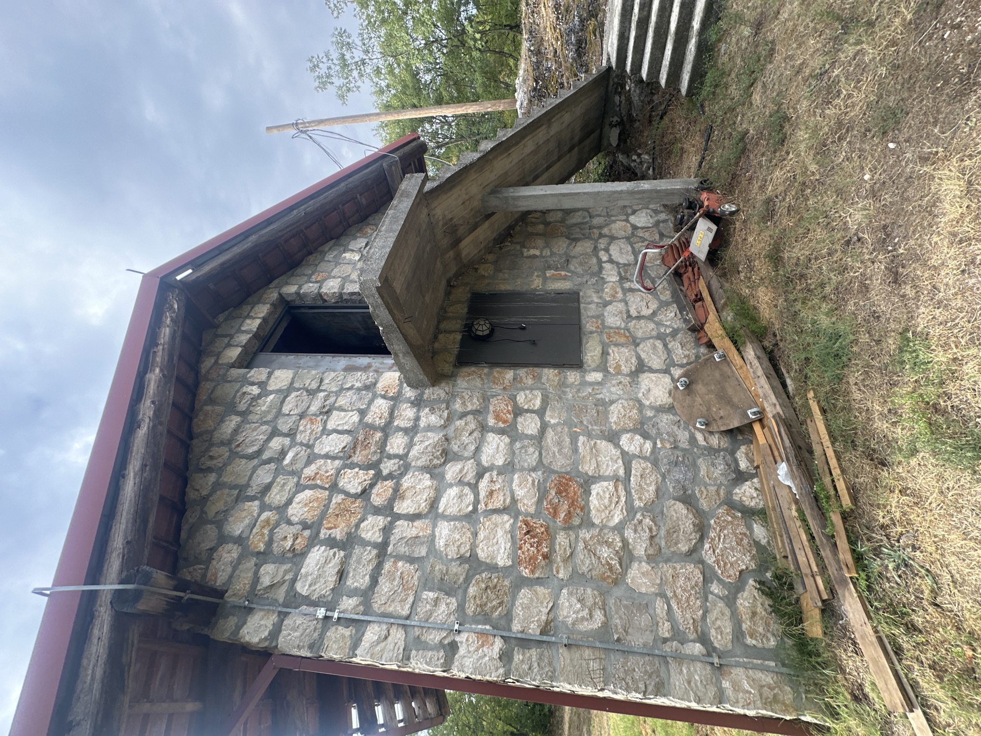 The image depicts the exterior of a small, stone-built structure in Cetinje, Montenegro. The building has a red-tiled roof with exposed wooden beams and a concrete overhang leading to a set of exterior stairs. The stone walls are constructed from irregular, rough-hewn stones of various sizes and colors, primarily light gray and tan with some reddish-brown accents, mortared together.
To the right of the building, a concrete staircase ascends alongside the structure. In front of the building, near the base of the stairs, there is a red and silver push lawnmower. A pile of wooden planks and a circular wooden object with metal fixtures are stacked near the lawnmower and against the stone wall. The ground surrounding the building is covered with dry grass and some scattered greenery. Trees and foliage are visible in the background, suggesting a semi-rural or wooded environment. The sky is overcast with heavy clouds. A wooden pole with electrical wires is situated to the right of the building.