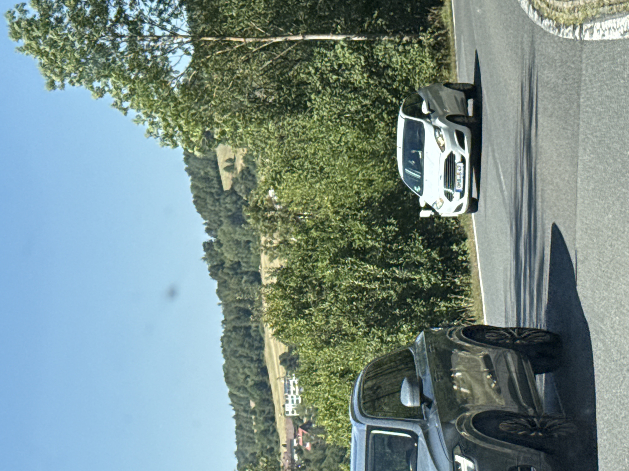 In Bad Lauterberg im Harz, Germany, on a sunny day with a clear blue sky, two vehicles are visible on a paved road winding through a lush, green landscape. On the left, a dark-colored SUV, possibly a Volvo XC90, is partially visible, showcasing its front left wheel and side mirror. To the right, a white Ford Fiesta is in motion, its front end clearly showing the grille, headlights, and license plate reading "OHA.B.47". The car appears to be turning on the road. The background is dominated by dense green trees and foliage, with a hint of buildings in the distance, suggesting a suburban or rural setting. The road surface is asphalt, with some tire marks visible. The overall scene depicts a typical drive through a scenic area.