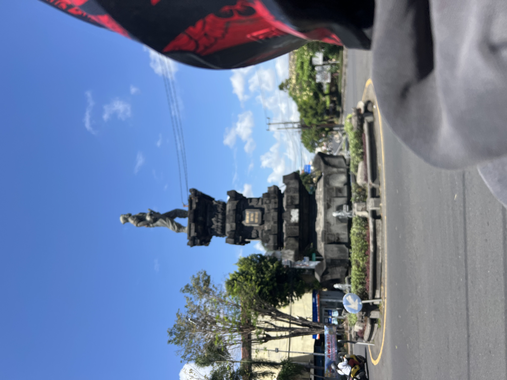 A statue of a man stands atop a tiered, stone monument at a traffic roundabout in Denpasar, Indonesia. The statue depicts the man in traditional clothing, with a beard and holding what appears to be a rifle. The monument is adorned with intricate carvings and has a fountain at its base. The roundabout is paved with asphalt and marked with yellow lines. Green foliage surrounds the monument. In the background, buildings and trees line the street. A motorcycle with a rider is visible on the left side of the roundabout, and a person's helmet, decorated with red patterns, is in the foreground on the right. The sky is blue with scattered white clouds.
