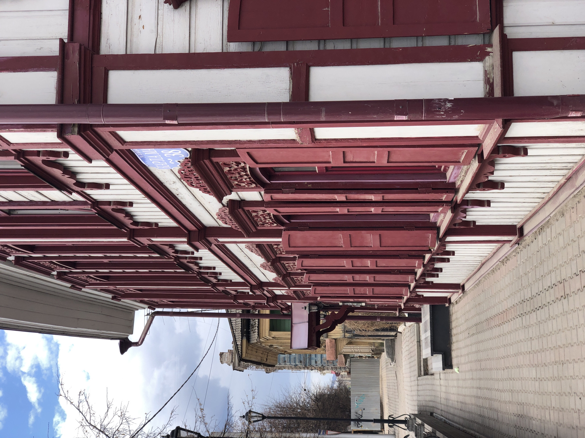 The image, rotated 90 degrees clockwise from its correct orientation, depicts an architectural street scene, likely in Krasnoyarsk, Russia. The primary subject is a long, covered wooden walkway or veranda, seen from underneath, attached to an older building.
The walkway structure is painted in a striking combination of dark red (maroon/burgundy) and white. It features intricate carved wooden details on the fascia boards and decorative supporting brackets, showcasing traditional craftsmanship. A dark red rain gutter runs along the edge of the roofline above the walkway. The building itself, partially visible above the structure, has white wooden siding.
The ground surface on the right appears to be a sidewalk, paved with light-colored interlocking bricks arranged in a patterned design. Looking down the street, several other buildings with varying historical architectural styles are visible, some featuring light yellow facades and distinct blue window frames. The streetscape also includes bare tree branches, utility poles with power lines, and a classic black street lamp. The sky above is partly cloudy with visible patches of blue.
No people or specific activities are visible, presenting a quiet, static urban environment. A notable detail is a blue address sign with white Cyrillic text, clearly reading "УЛИЦА МИРА 28" (Ulitsa Mira 28), which translates to "Peace Street 28". This sign is affixed to the underside of the walkway structure, providing a specific location cue for the city. The overall impression is that of a historic urban corridor captured on a daytime, partly cloudy day.