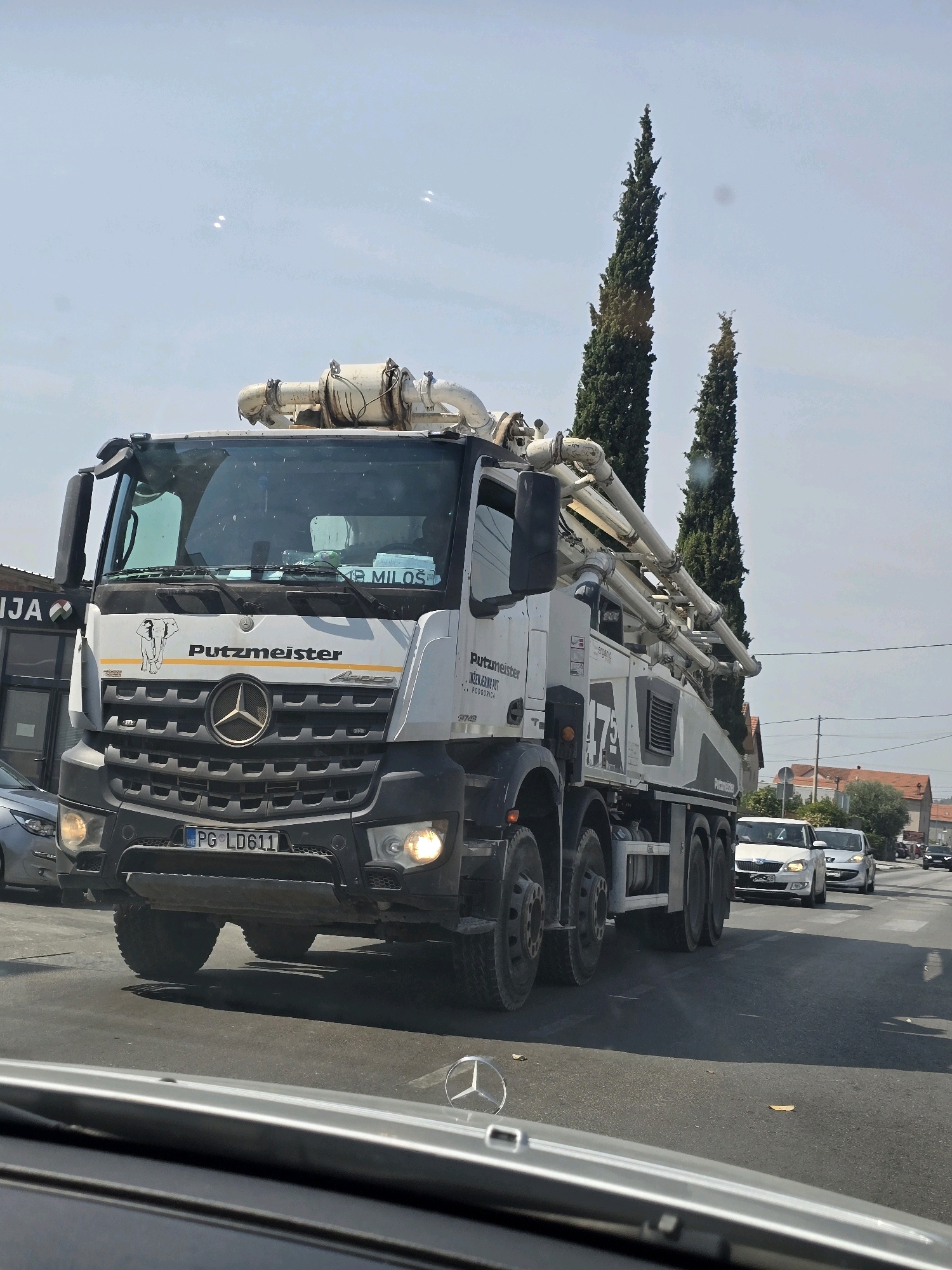 The image shows a white Mercedes-Benz concrete pump truck with the "Putzmeister" logo and "Arocs" model designation on the front. The license plate reads "PG LD611," indicating it is registered in Podgorica, Montenegro. The truck is in motion on a road, possibly in a commercial or industrial area, as suggested by the building on the left with the partially visible "NJA" logo. Two tall cypress trees are visible behind the truck. In the background, there are other vehicles on the road and buildings with red tile roofs. The sky is bright and clear, suggesting a sunny day. The perspective is from inside another vehicle, looking forward towards the truck. There are no visible people in the image.