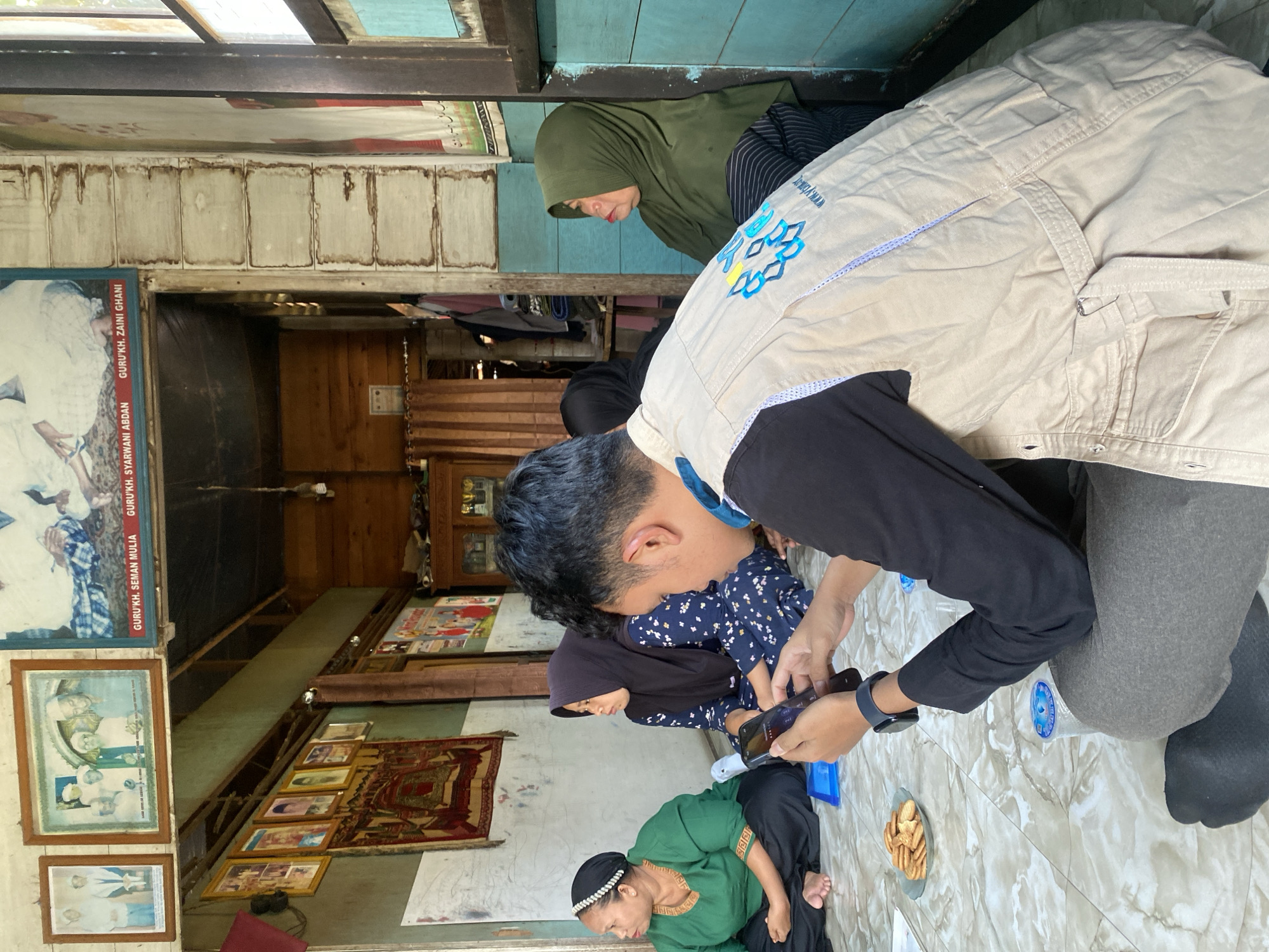 In the city of Bawahan Selan, Indonesia, a group of people are gathered indoors. In the foreground, a young man wearing a beige vest over a black shirt is holding a smartphone. He is seated on the floor, facing away from the camera, with his legs crossed. To his left, a woman in a green shirt and headscarf is also seated on the floor, looking towards the left. Next to her is another woman wearing a dark blue patterned dress and a black hijab, looking towards the right. Further to the right, a woman in a green hijab is seated, leaning against a wall. The floor is tiled with a marble-like pattern. On the floor, there is a plate of crackers and a cup of water. The background shows a room with wooden walls and a low ceiling. Several framed pictures and a rug are visible on the walls. The overall environment suggests a casual indoor gathering in what appears to be a home or community space.