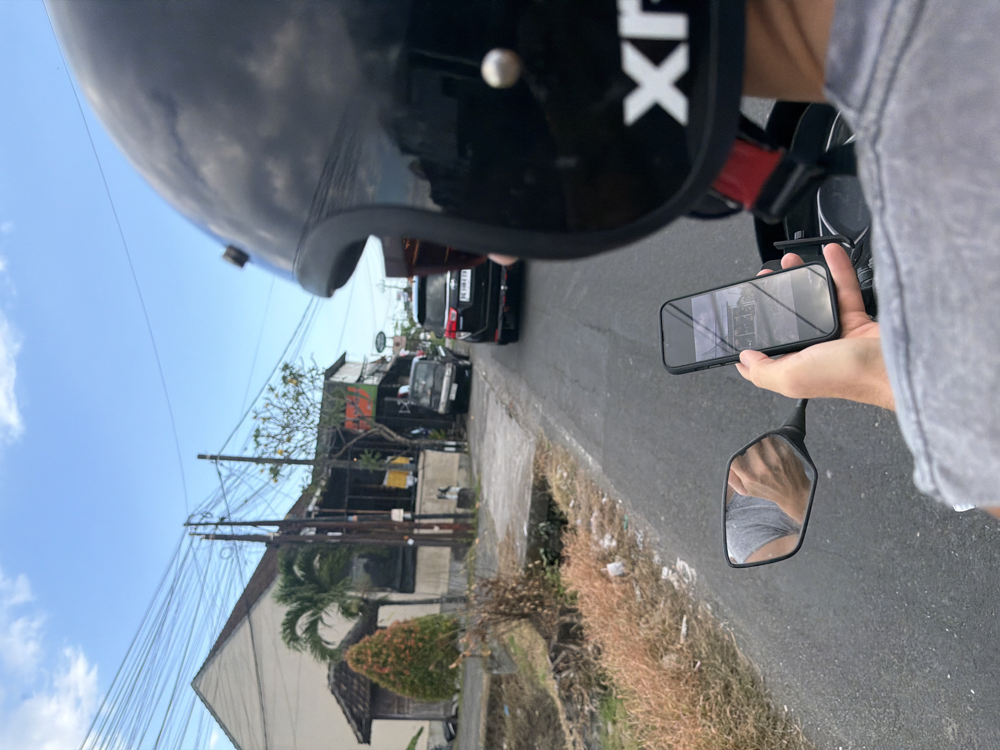 A first-person perspective image from a rider on a motorbike in North Kuta, Indonesia. The rider is wearing a black helmet, partially visible in the foreground. The rider holds a smartphone in their right hand, apparently displaying a photo or video. A motorcycle mirror reflects the rider's arm and grey shirt.
The background shows a narrow street with a black SUV and a smaller vehicle further ahead. Buildings are visible on the side of the road, obscured by power lines. The sky is partially cloudy. The roadside has dry grass and debris.
