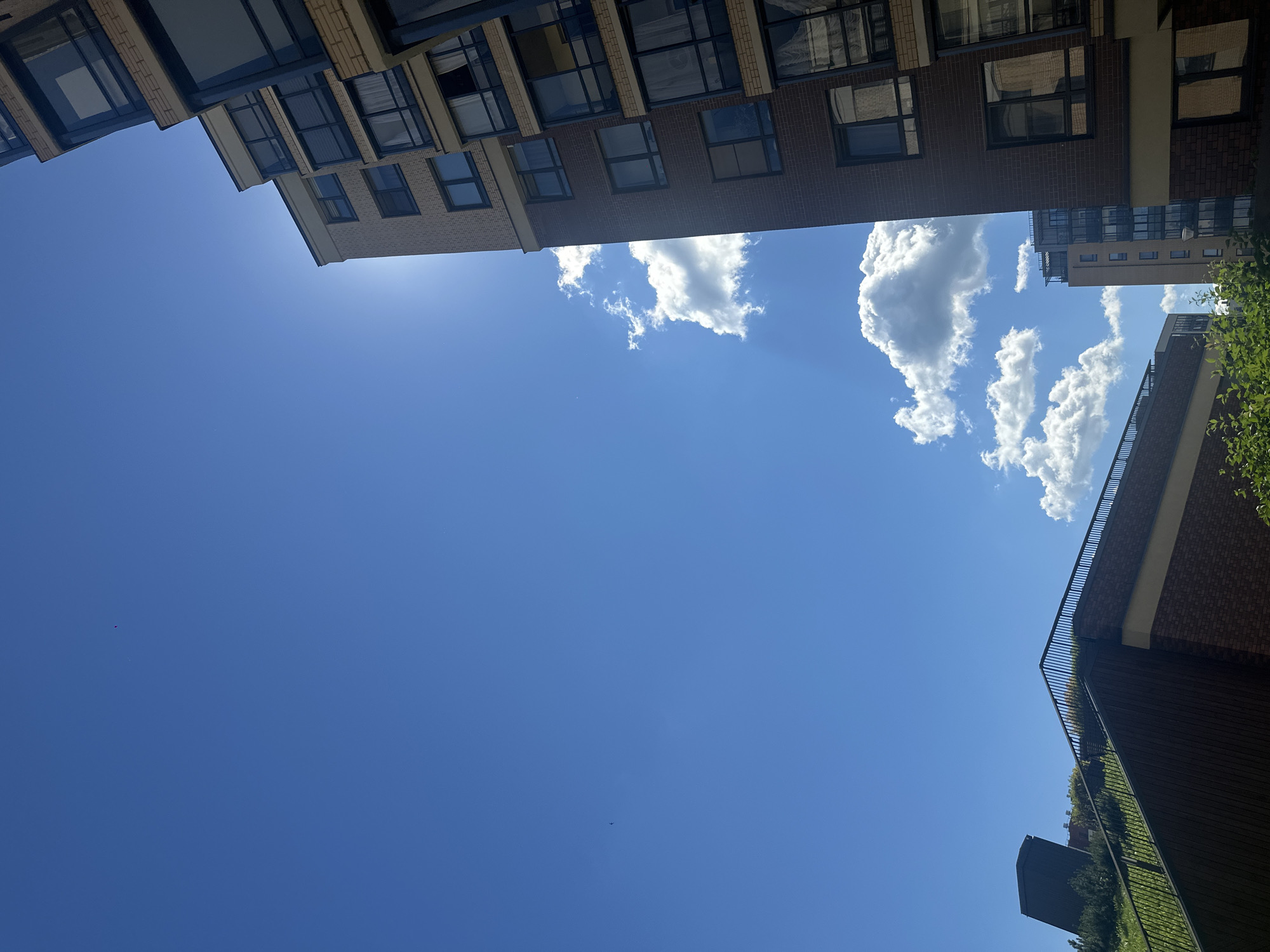The image captures a low-angle view of an urban environment under a clear blue sky with scattered clouds. On the right side of the frame, a modern brick apartment building with multiple floors and dark-framed windows dominates the scene. The sun is visible, creating a lens flare effect as it peeks from behind the building. In the foreground and left side of the image, a sloped green roof with a black railing and brick facade is visible, likely part of another building or structure. Further in the background, another apartment building with a lighter facade can be seen. The overall impression is a contemporary cityscape on a bright, sunny day.
