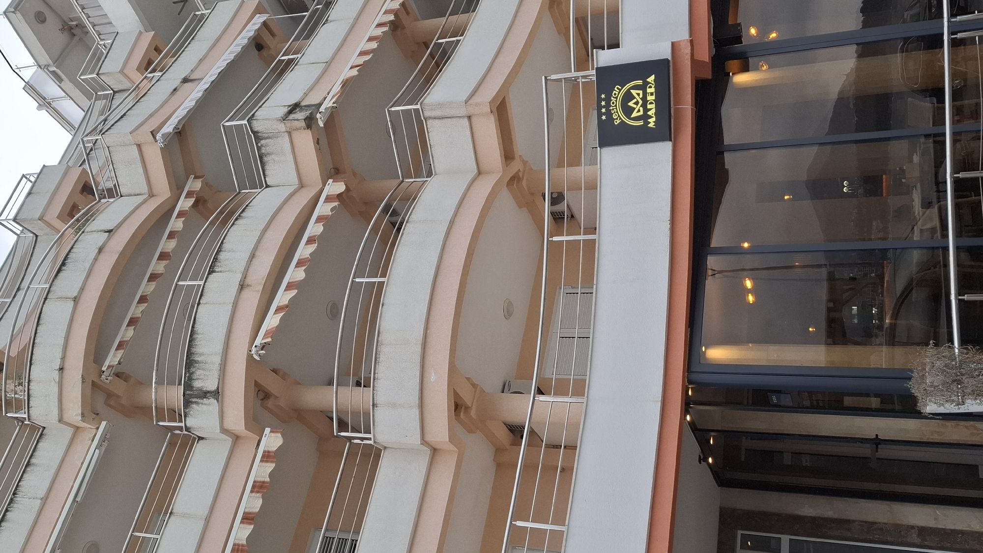 The image displays a multi-story building located in Boreti, Montenegro, under an overcast sky. The structure features five visible floors, each with curved balconies adorned with silver-colored metal railings. The building's exterior consists of peach-toned support columns and balcony undersides, contrasted with light grey or white balcony facades. Several balconies are equipped with retractable striped awnings in white and peach/orange, some partially extended. Visible grime and dark streaking are present on the undersides of multiple balconies.
The ground floor houses a commercial establishment identified by a black rectangular sign with gold lettering that reads "Restoran MADERA" and displays a four-star rating. The restaurant's facade is composed of large glass panels, reflecting a distant mountainous landscape, scattered streetlights, and a partial view of a vehicle. Interior lighting is visible through these reflections. Air conditioning units are mounted on the exterior walls of some upper-floor balconies. No individuals are present in the scene.