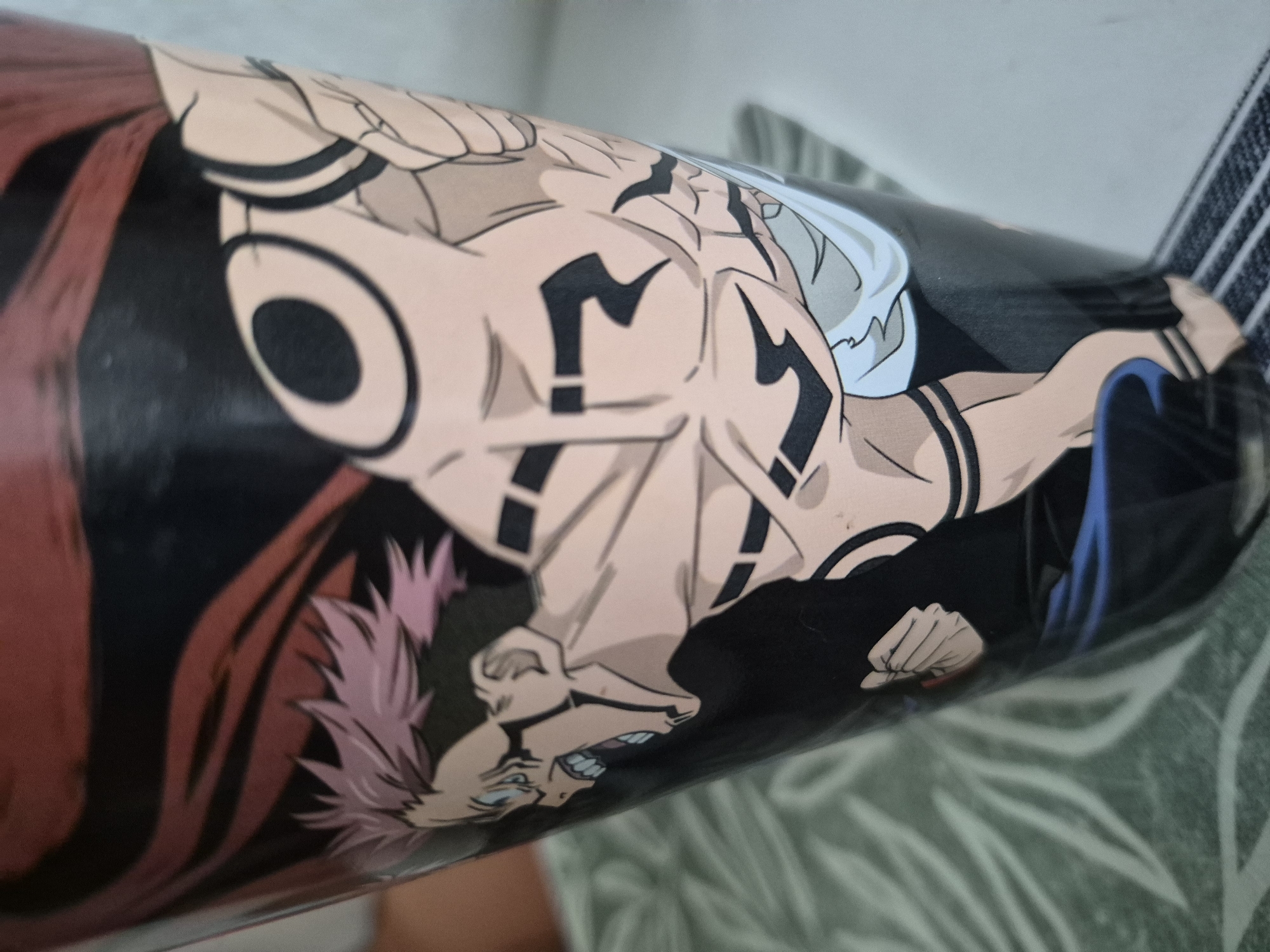 The image shows a close-up of a black cylindrical object, likely a cup or container, decorated with an anime character. The character, depicted in a dynamic pose with pink spiky hair and visible muscles, is a central focus. The character has blue eyes and an open mouth, suggesting an action or expression of shouting. Intricate black markings, resembling tattoos or symbols, are prominently displayed on the character's arms and torso. The background of the object features dark red and black abstract shapes. The object itself is placed on a surface with a green, leaf-patterned texture, and a grey striped mat or floor is visible at the bottom. The lighting suggests an indoor environment. No specific city or location details are discernible.
