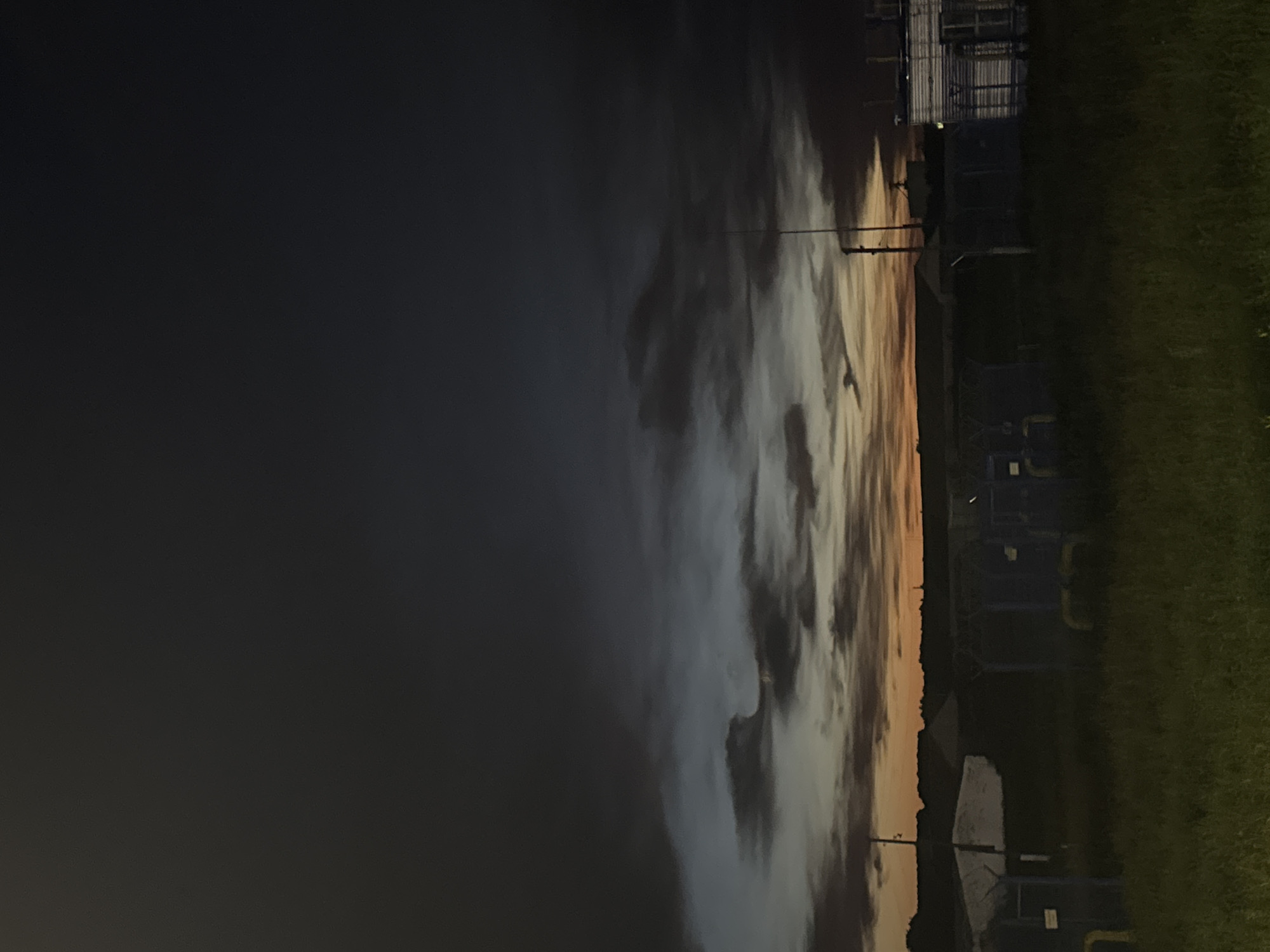 The image depicts a dimly lit outdoor scene at dusk, likely in the vicinity of Анатыш, Russia, given the context. The sky is a dramatic display of dark, ominous clouds, with streaks of orange and yellow near the horizon, suggesting a sunset or the remnants of it. In the foreground, there is a grassy field with some sparse vegetation. Scattered throughout the midground are various structures, including what appear to be industrial or utility buildings with metallic exteriors and fencing. There are also what seem to be yellow pipes or conduits visible. A thin, tall pole, possibly an antenna or lighting fixture, stands prominently in the middle ground. The overall atmosphere is one of quiet desolation and a transition into night. There are no people or animals visible in the image. The scene suggests an industrial or agricultural area on the outskirts of a settlement.