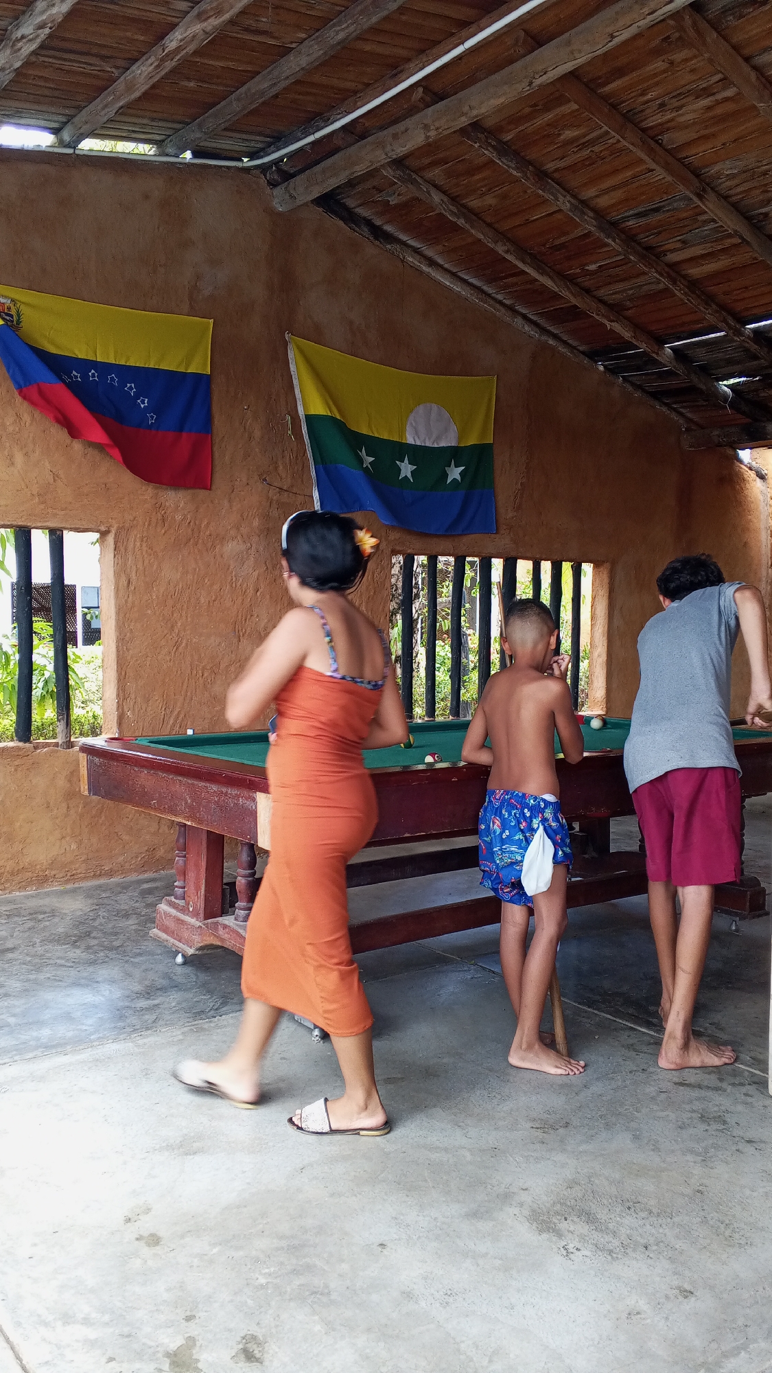 The image depicts three individuals near a pool table under a thatched roof structure. Two flags are mounted on the wall behind the pool table. The individual on the left is wearing an orange dress and sandals. The middle person, a child, is shirtless and wearing blue patterned shorts. The person on the right is wearing a gray shirt and maroon shorts. The pool table is green, and several pool balls are visible. The walls are textured and painted in an earthy tone. The exact location and city are unknown.