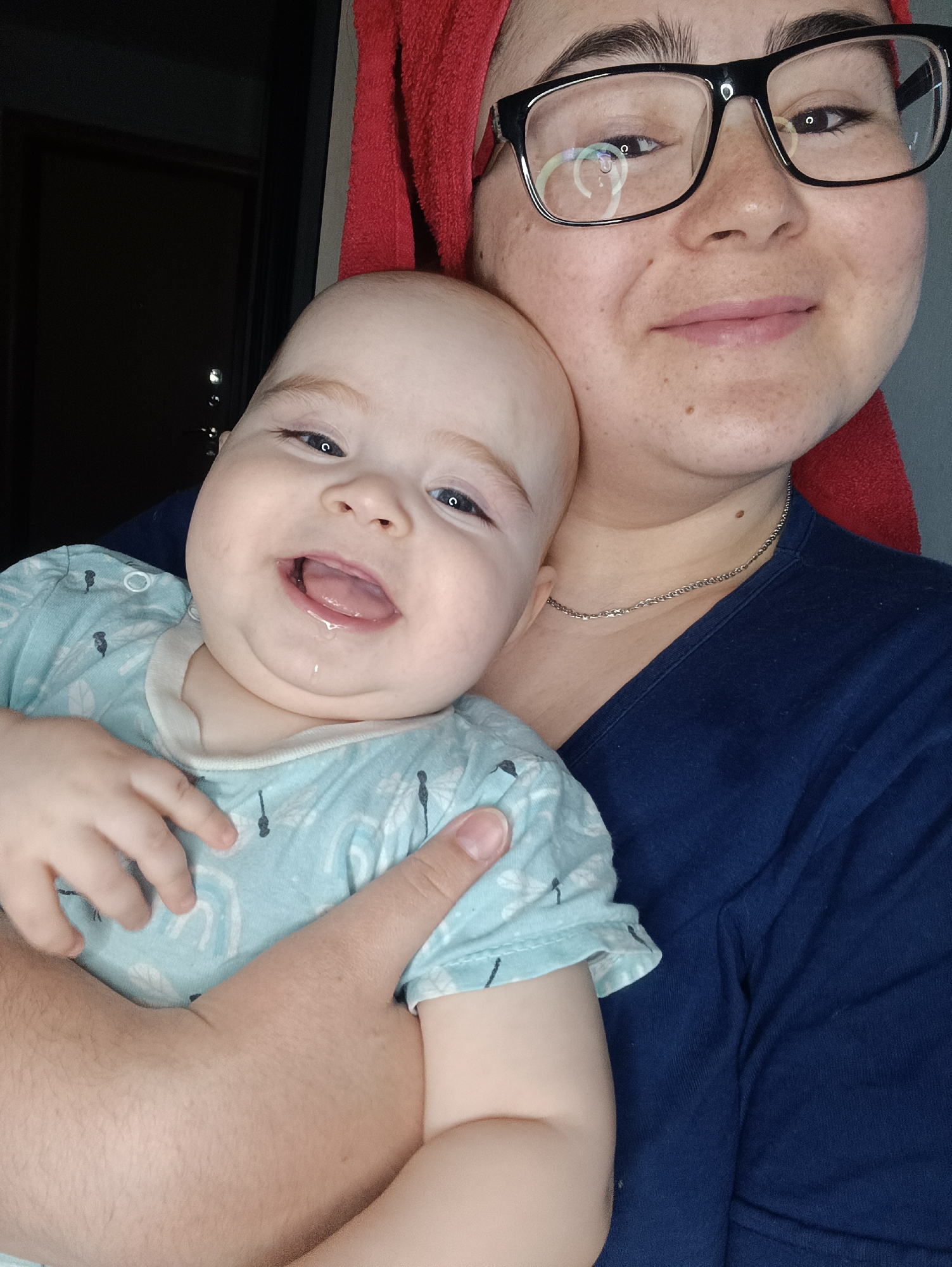 A medium shot depicts a woman holding a baby, indoors. The woman, likely in her late twenties or early thirties, wears black-framed glasses and a red towel is wrapped around her head. She has fair skin and freckles and is wearing a dark blue top. She is also wearing a silver chain necklace. The baby, held in her arms, is wearing a light blue patterned shirt and has a droplet of saliva escaping their open mouth. The baby is fair-skinned and appears to be around six to twelve months old. The baby has a happy expression on their face, and is looking at the camera. A hand is wrapped around the baby's torso, presumably the woman holding the baby. Behind them, is a dark doorway. The scene is lit by artificial light, and the location is listed as Noviy, Russia. No landmarks or details confirm the city.