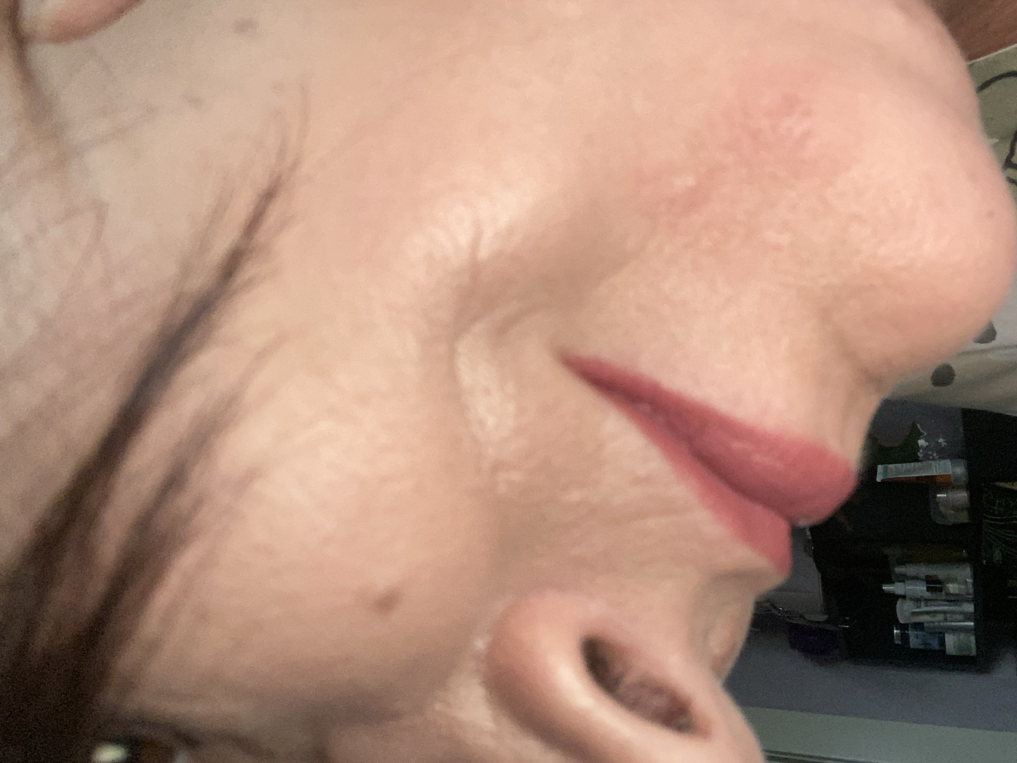 The image is a close-up, angled shot of a person's face, focusing on the nose and lips. The skin appears to have some texture and possibly makeup on the lips, which are a reddish-pink color. The background is out of focus and shows a shelf with various bottles and containers, suggesting an indoor setting, possibly a bathroom or vanity area. There is no indication of the city of Novokuznetsk, Russia, in the image.