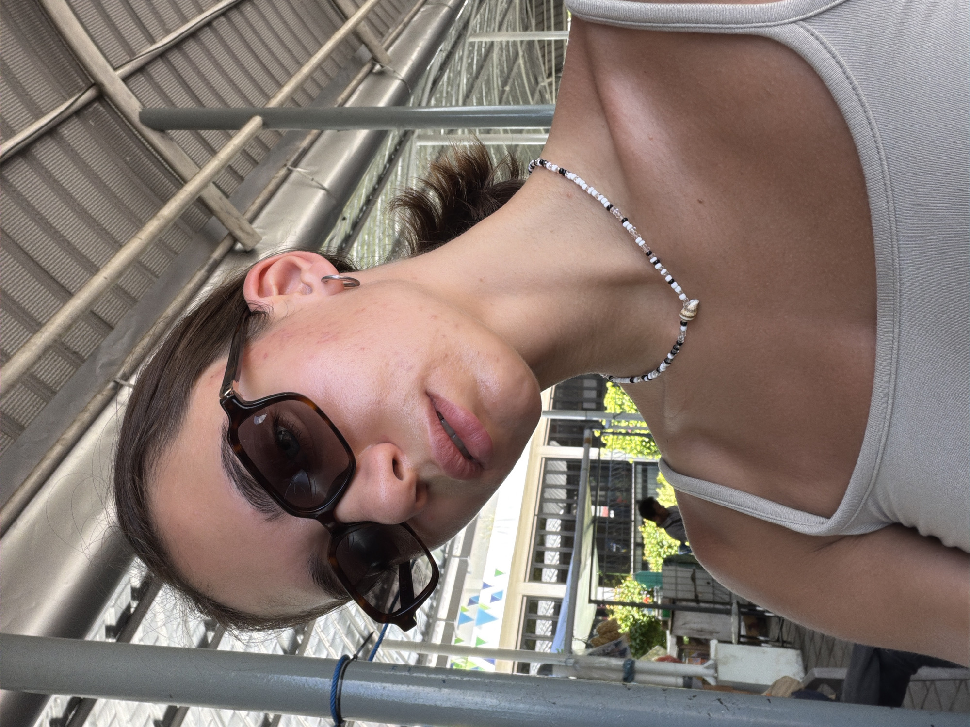 A woman wearing sunglasses and a white tank top is facing the camera while standing outdoors. She has a beaded necklace and an earring. The background shows a building with windows, trees, and various objects, suggesting a market or a public area. The location appears to be in the city of Gunung Malang, Indonesia, based on the environment. Metal structures are visible in the foreground, possibly part of the surrounding architecture or infrastructure.