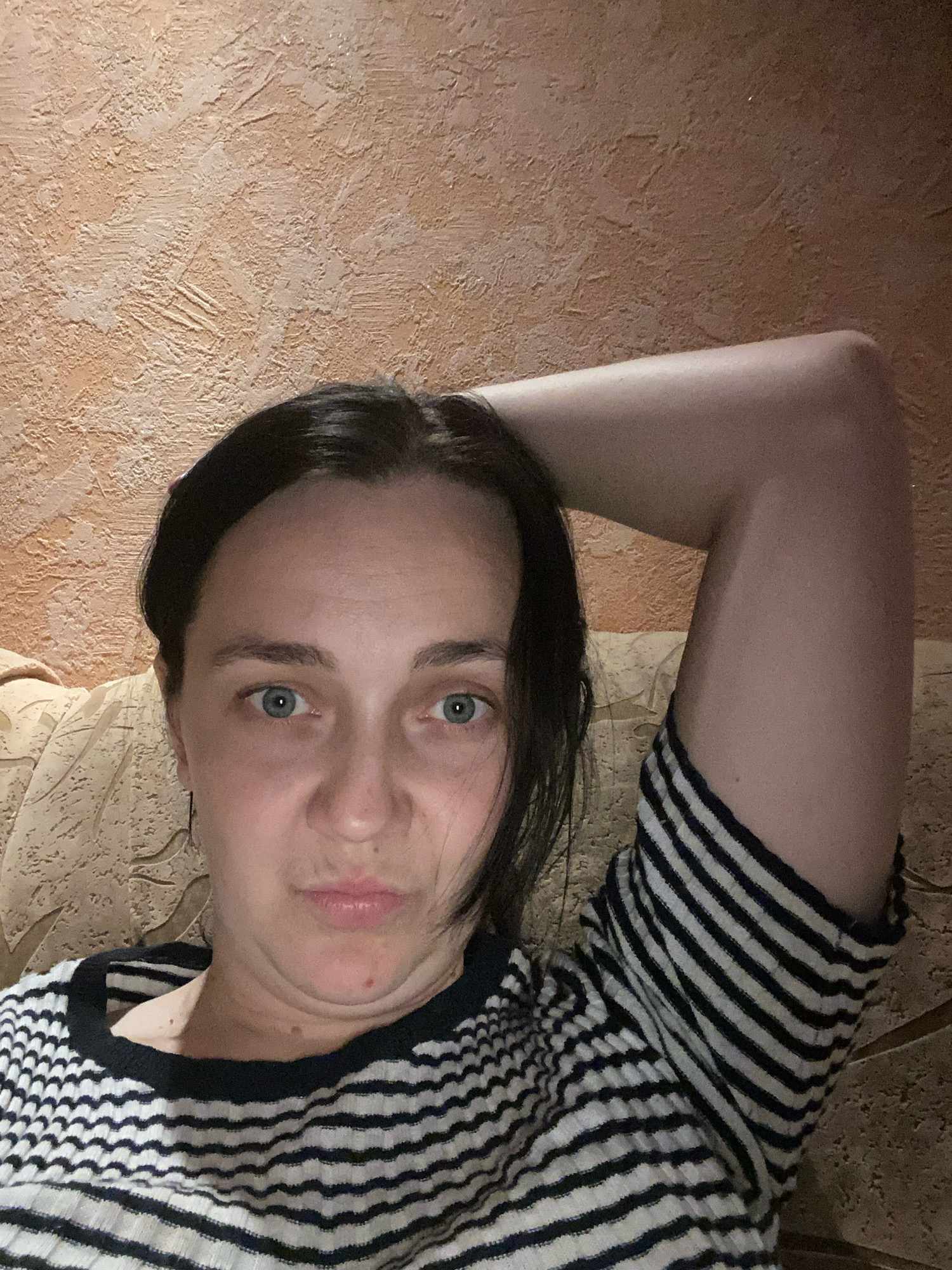 A woman is lying down on a textured couch, with her arm raised behind her head. She is wearing a black and white striped sweater. The background is a textured orange wall. The lighting is dim, suggesting it is nighttime or indoors.