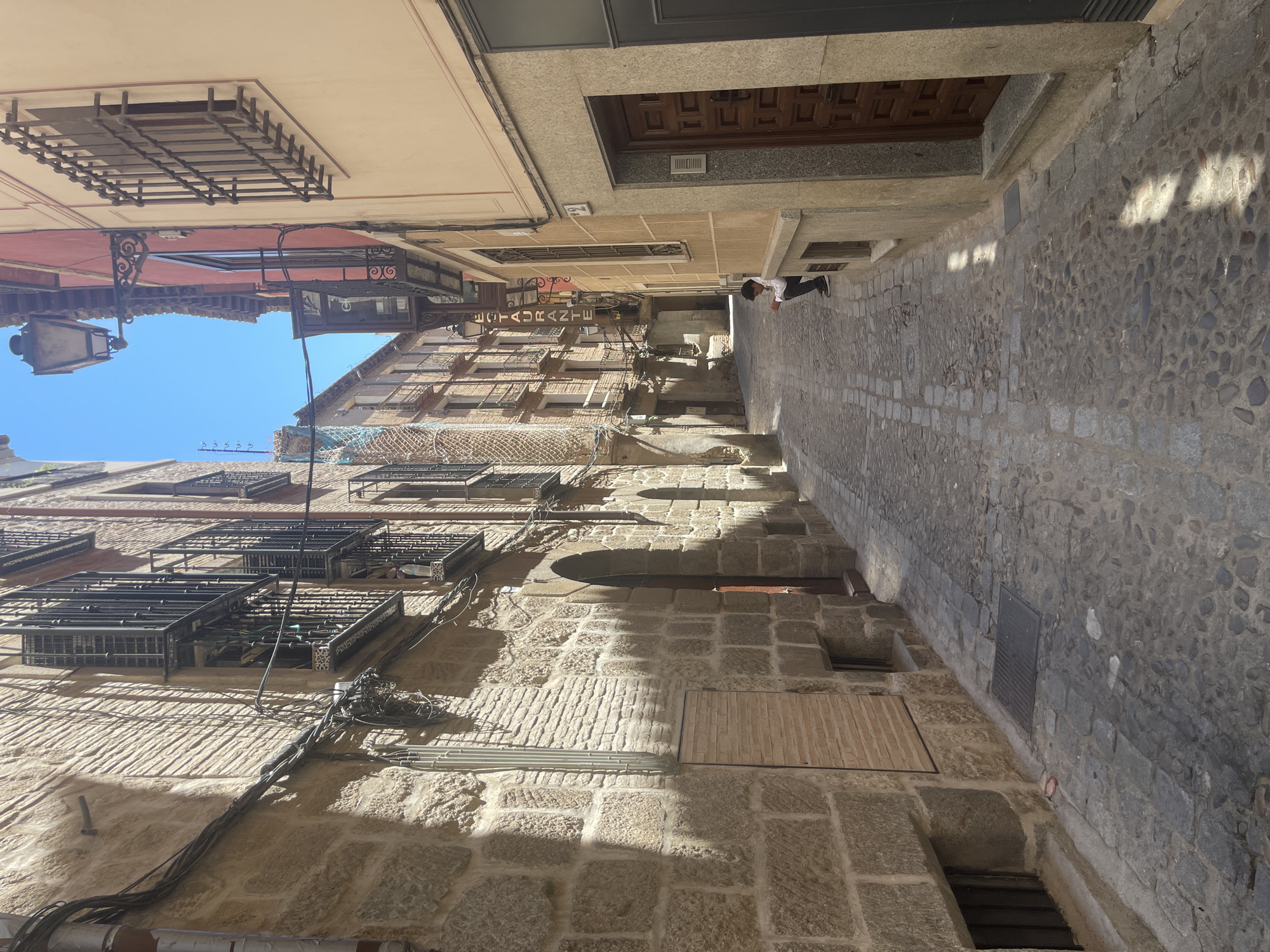 The image depicts a narrow cobblestone street in the historic district of Toledo, Spain. The buildings lining the street are constructed with stone and brick, featuring wrought-iron window grates and balconies. Some of the buildings have a weathered appearance, suggesting their age. The street is illuminated by sunlight, casting distinct shadows on the ground and buildings. A lone individual, a young person wearing a white shirt and dark pants, is seated on a low ledge on the right side of the street, looking down at the ground. The street appears quiet and somewhat deserted. Overhead, a vintage-style street lamp is visible. The architecture suggests a location within a Mediterranean or European city, consistent with Toledo's historical character.
