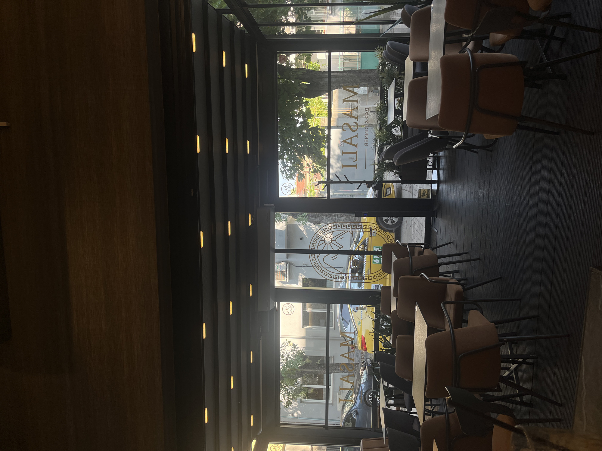 The image depicts the interior of a restaurant or cafe, likely located in Plovdiv, Bulgaria, as suggested by the signage visible through the glass windows. The establishment features a modern, minimalist design with dark wooden flooring and a ceiling with embedded lighting. Numerous tables and chairs, upholstered in brown and grey fabrics, are arranged in the dining area. Large glass windows offer a view of the street outside, where cars, including a yellow taxi, are parked. The exterior also shows buildings and trees, indicating an urban setting. The signage on the windows appears to be in a foreign language, possibly Cyrillic, and includes the name "MASALI BAR & DINNER". The lighting suggests it is either early morning or late evening. No people are visible in the immediate foreground or at the tables. The overall ambiance is calm and unoccupied.