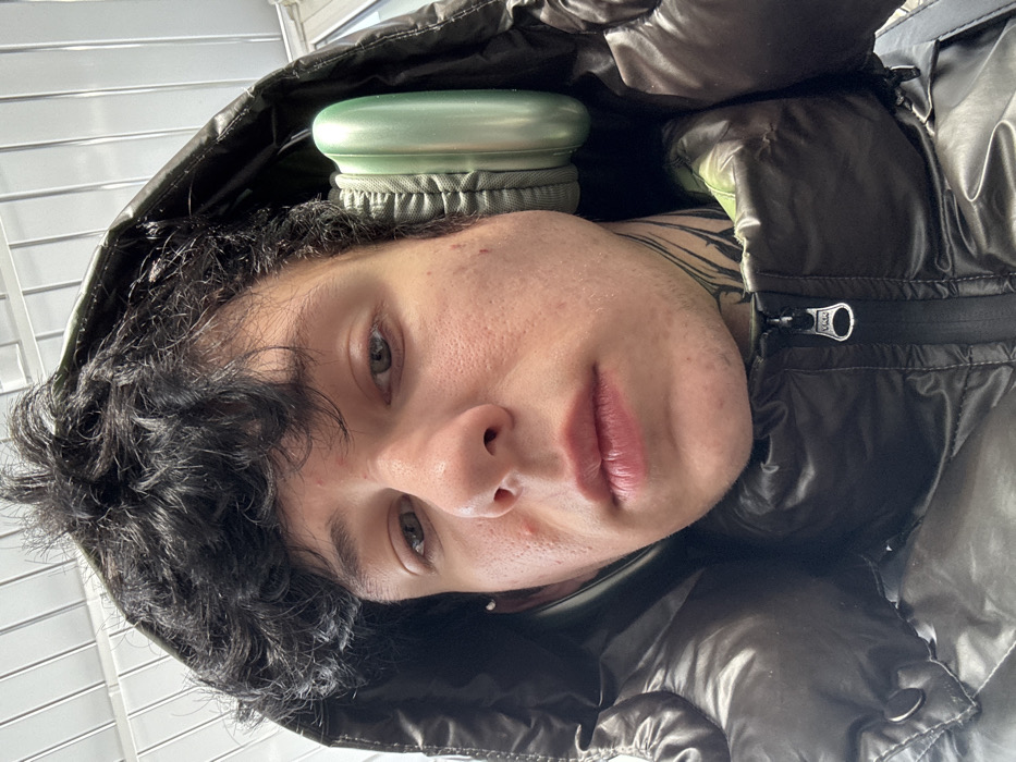 A close-up, eye-level portrait captures a young male subject in Tatishchevo, Russia. The individual, appearing to be in his late teens or early twenties, has dark, curly hair protruding from beneath a dark, glossy puffer jacket hood. His skin is fair, with visible blemishes on the cheeks and forehead. He wears light green over-ear headphones, partially obscured by the hood. Black ink tattoos are visible on the left side of his neck, and a small silver stud earring is present in his left earlobe. The subject maintains a neutral expression, with slightly parted lips, and looks directly at the camera. The background above his head features a light-colored, ridged or slatted structure, indicative of an indoor or covered architectural setting.