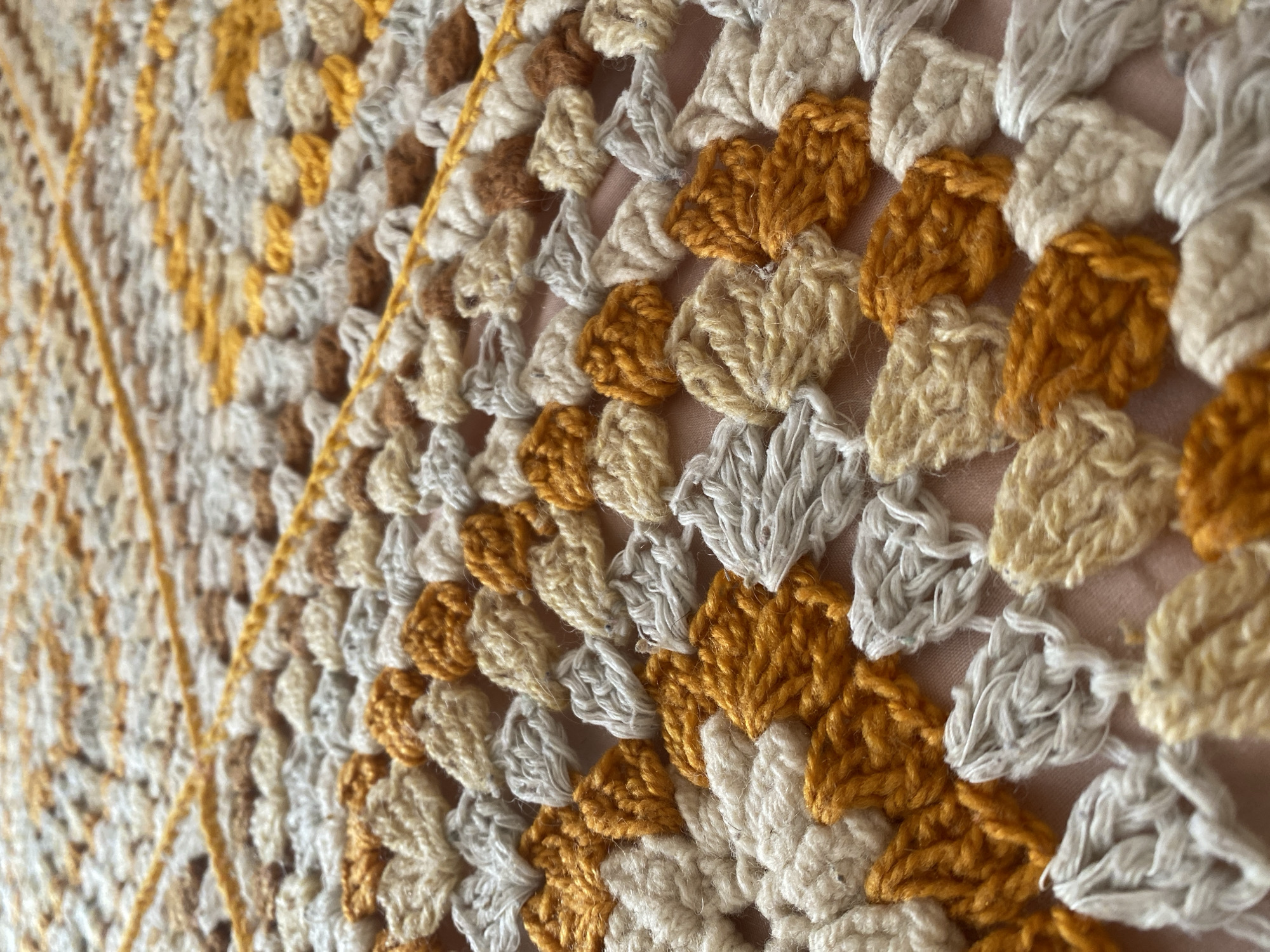 This is a close-up, eye-level shot of a crocheted blanket or textile. The textile features a repeating pattern of clusters of stitches in various earth tones, including cream, light beige, rust brown, and a golden yellow. There are also strands of a bright yellow yarn woven across the surface of the textile, creating a linear design element. The stitches are densely packed, giving the fabric a textured appearance. The background appears to be a solid, light-colored surface, possibly a bed or table, also in a light pinkish hue. The image is focused on the texture and pattern of the crocheted material, with no indication of location in Anos, Russia, or any people, actions, or interactions.