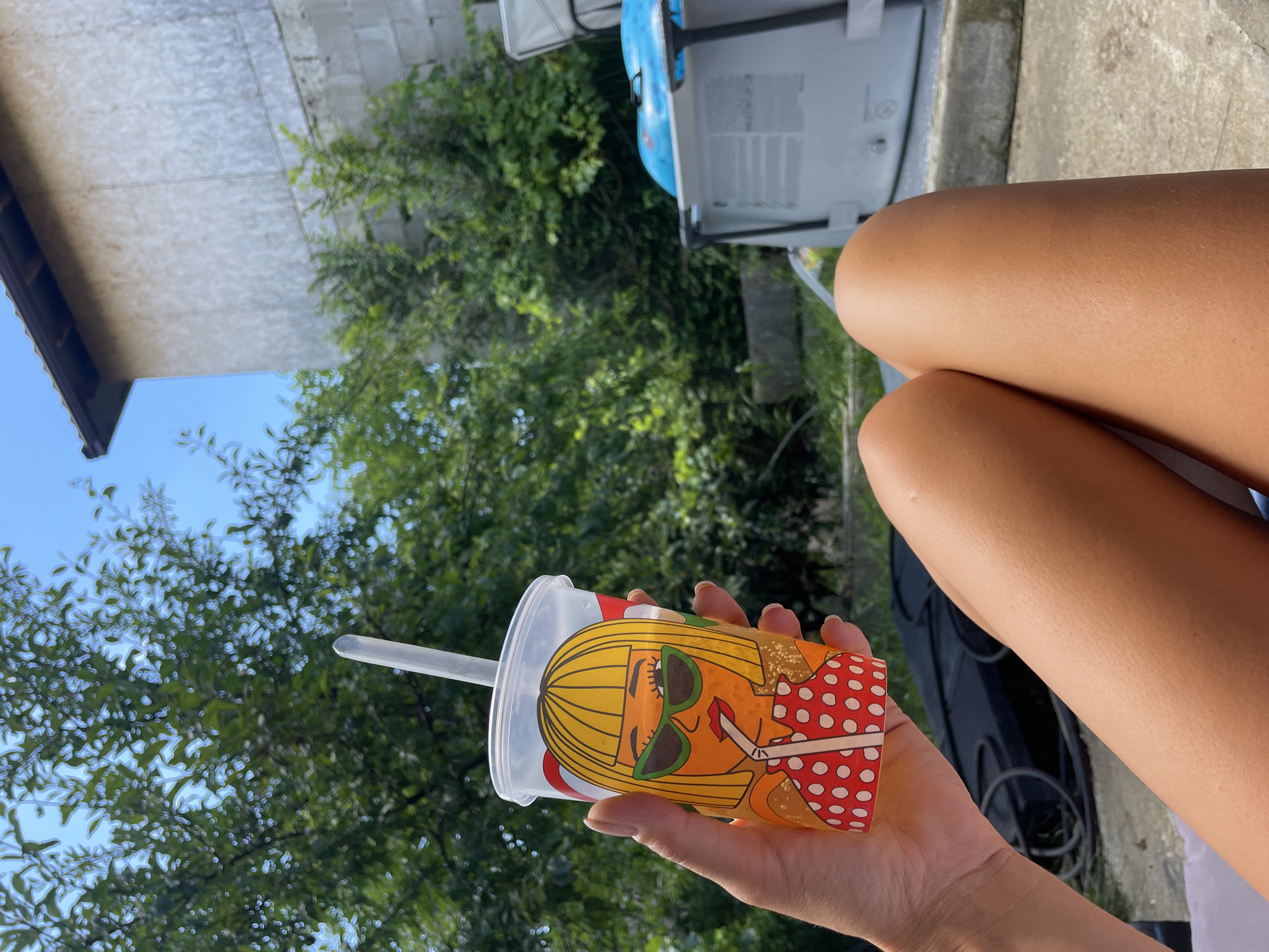 A person's tanned legs are positioned in the foreground, with a person's hand holding a transparent plastic cup in the upper left quadrant. The cup features a cartoon illustration of a woman with blonde hair, sunglasses, and a red and white polka dot top, drinking from a straw. Bubbles are visible within the beverage inside the cup. The background consists of lush green trees, a clear blue sky, and a grey cinder block building with a brown roof. A blue inflatable pool can be seen to the right of the frame. The scene appears to be outdoors in a residential backyard, likely during daylight hours. The location is not Baranovka, Russia.