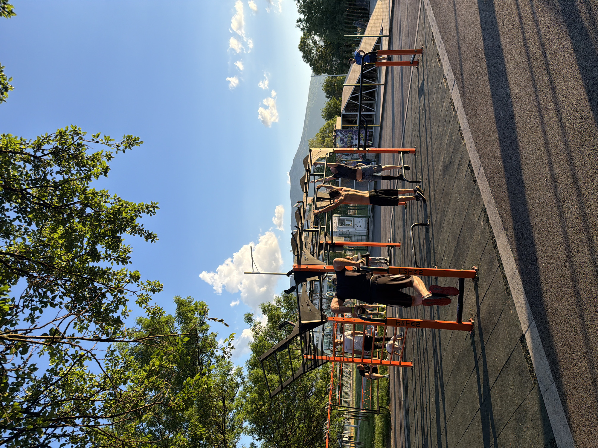 Outdoors in Sofia, Bulgaria, a group of five men are exercising on parallel bars in a park. The bars are orange, and the ground beneath them is black. To the left of the frame are trees, and in the background, a mountain range and a blue sky with clouds are visible. One of the men wears a black shirt and black shorts, another is shirtless, and another wears a blue shirt. A gray asphalt path runs along the right edge of the scene.