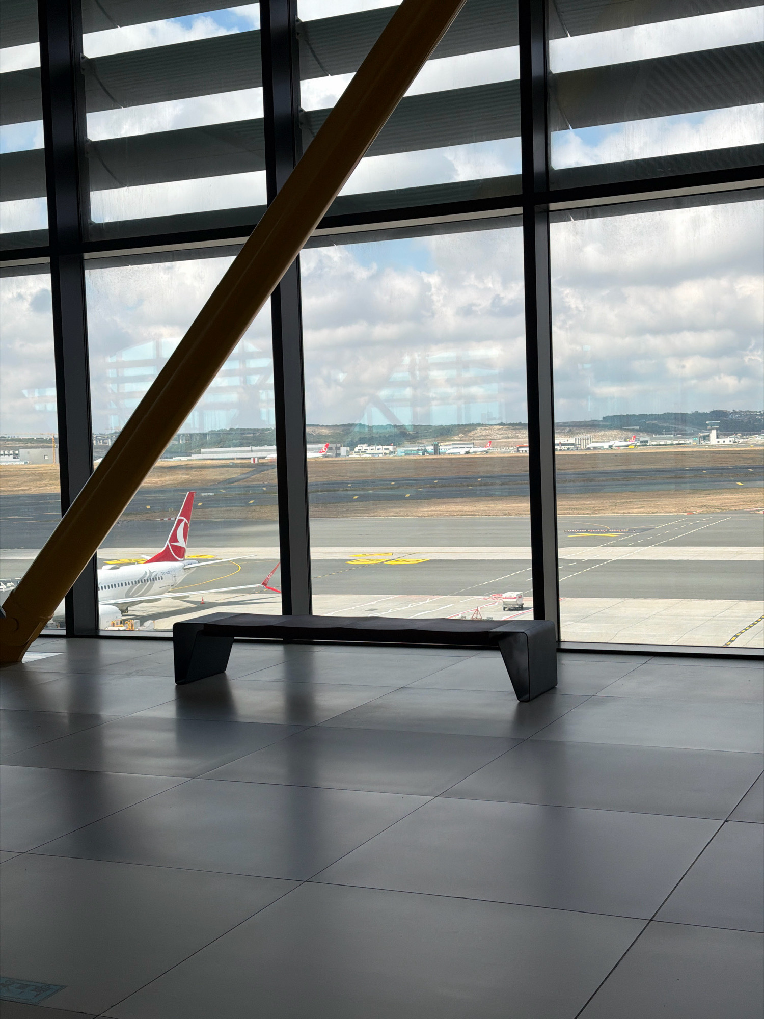 The image depicts a modern airport terminal interior with a large window overlooking an airfield. A dark, angular bench is positioned in the foreground, casting a shadow on the grey tiled floor. Through the window, several airplanes are visible on the tarmac, including a Turkish Airlines plane with its distinctive red and white logo. The airfield is lined with runways and taxiways, and in the distance, there are airport buildings and control towers. The sky is partly cloudy, with a mix of blue sky and white clouds. The architecture of the terminal is characterized by large glass panes and a prominent yellow diagonal support beam. The location is identified as an airport in Arnavutköy, Turkey, likely Istanbul Airport, given the presence of Turkish Airlines aircraft and the common architectural style of modern international airports. The scene is static and devoid of people, suggesting a quiet moment at the airport.