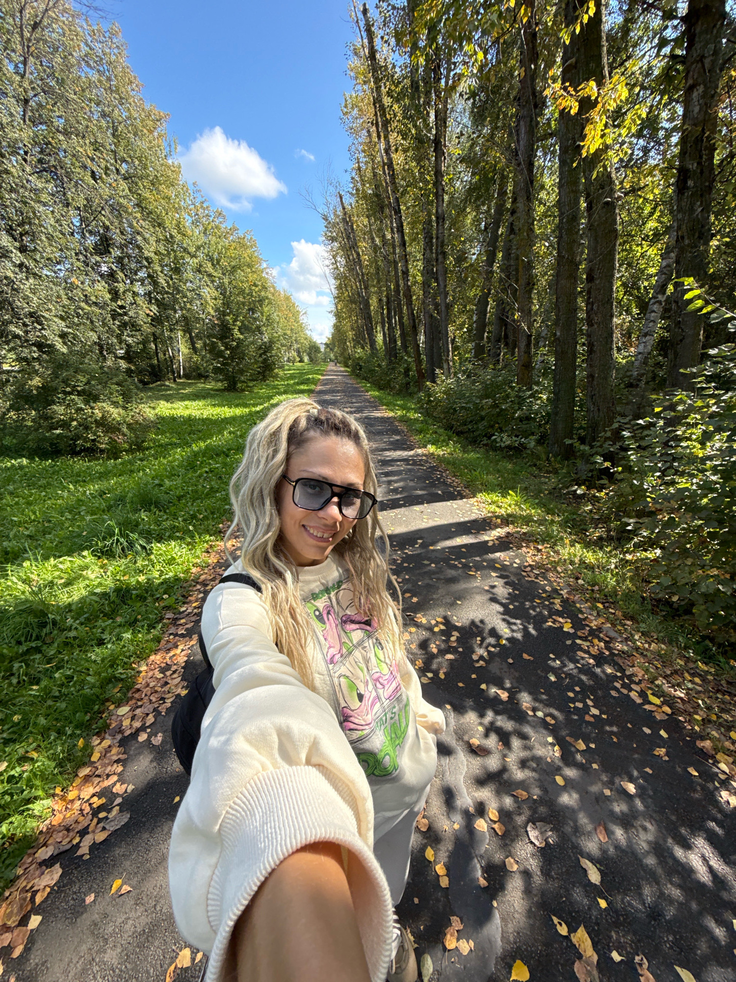 A woman with blonde hair tied in a high ponytail and large black sunglasses takes a selfie while walking on a paved path in Rusanovo, Russia. She is wearing a light-colored sweatshirt with a cartoon graphic and grey pants. A black backpack is visible on her shoulders. The path is lined with trees and green grass, and fallen yellow leaves are scattered on the ground. The sky is blue with scattered white clouds. The sun casts shadows on the path, indicating it is a sunny day.