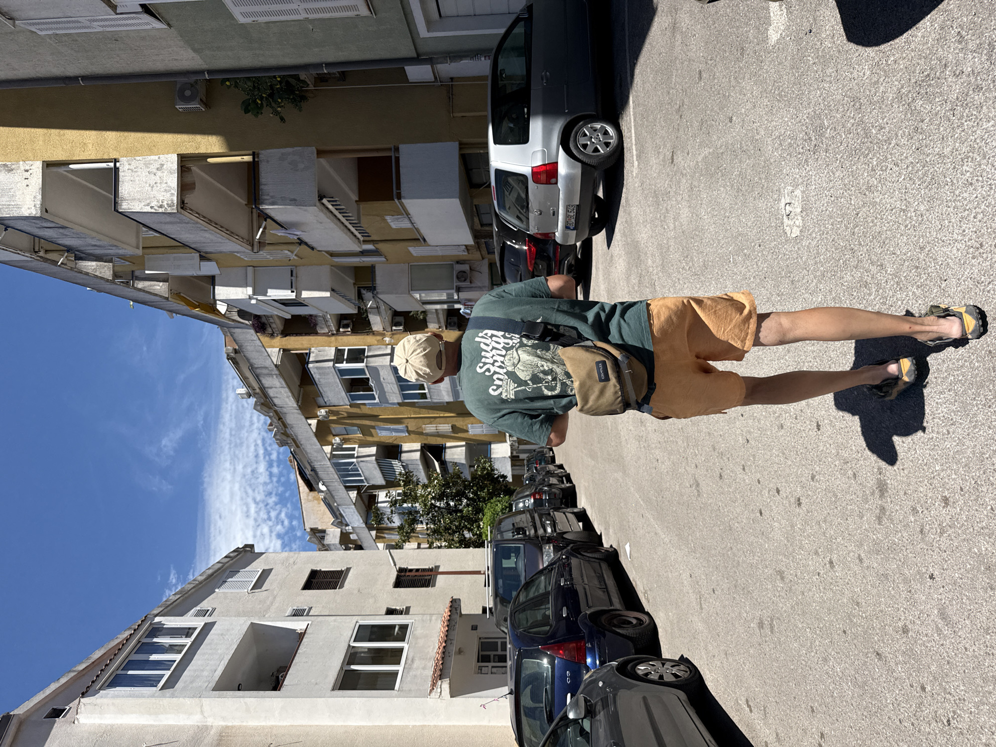 The image shows a person walking away from the camera down a narrow street lined with apartment buildings and parked cars. The person is wearing a green t-shirt with a graphic print, orange shorts, a beige baseball cap, and sandals. They have a brown crossbody bag slung over their shoulder. The buildings are typical of a residential area, with balconies and air conditioning units visible. The street is paved and appears to be in good condition. The sky is blue with scattered white clouds. The scene suggests a warm, sunny day in a European city.