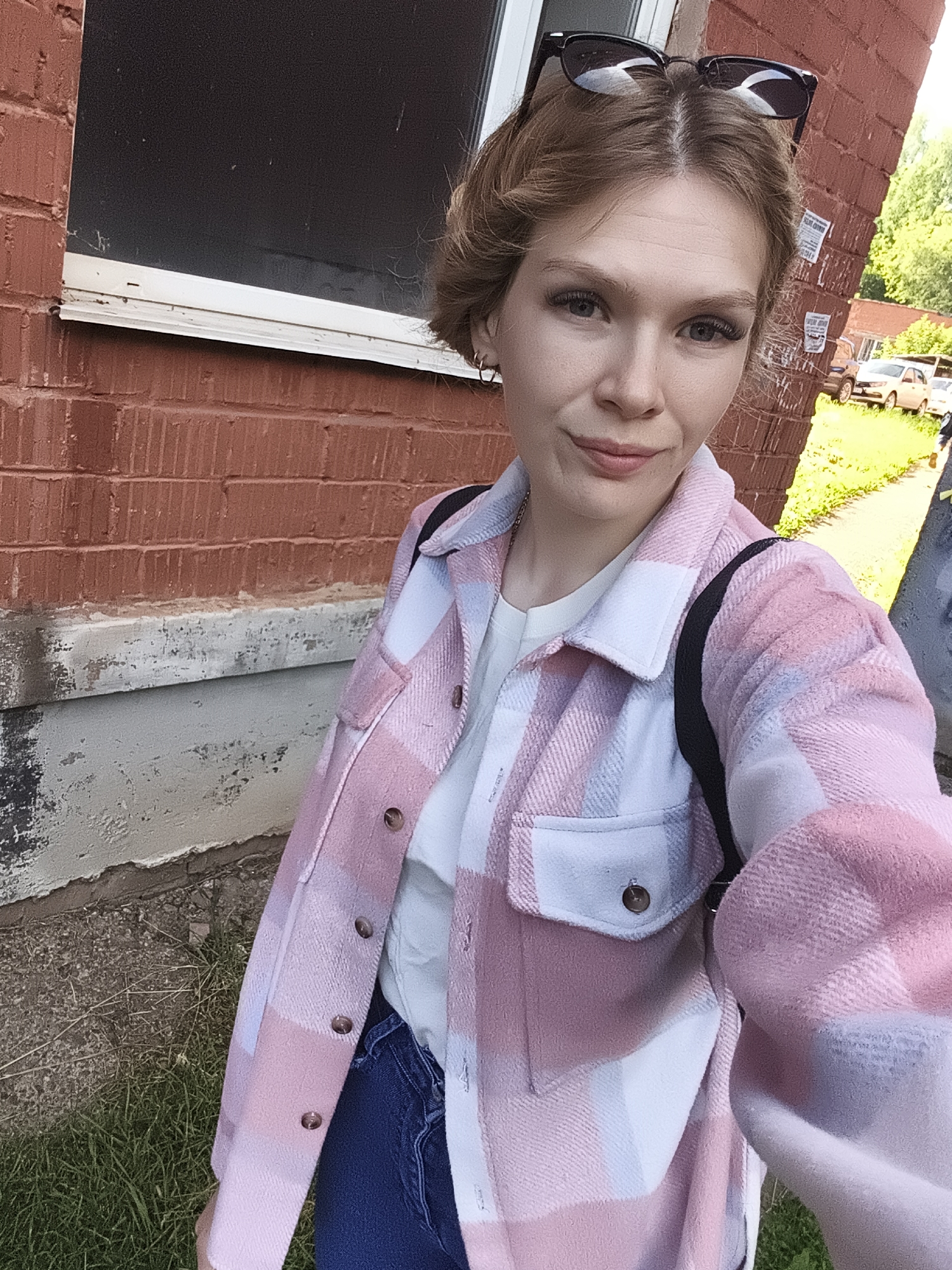 A young Caucasian woman with short, light brown hair and sunglasses perched on her head stands in front of a red brick building. She is wearing a pink and white plaid shirt jacket over a white t-shirt and blue jeans. A black backpack is slung over her shoulder. The background shows a window on the brick building and a grassy area with some cars parked in the distance. The scene appears to be in Izhevsk, Russia, given the context provided by the user.