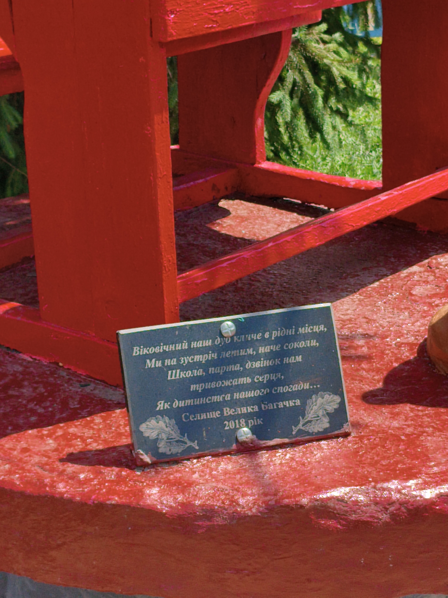 A dark blue plaque with white text is affixed to a bright red, possibly painted, wooden structure. The text on the plaque appears to be in Ukrainian and reads: "Віковічний наш дуб кличе в рідні місця, Ми на зустріч летим, наче соколи, Школа, парта, дзвінок нам тривожать серця, Як дитинства нашого спогади... Селище Велика Багачка 2018 рік". This translates to: "Our eternal oak calls to native places, We fly to meet, like falcons, School, desk, bell disturb our hearts, Like our childhood memories... Village of Velyka Bahachka, 2018." The structure itself is painted a vivid red and is situated outdoors, with blurred green foliage visible in the background, suggesting a natural or park-like setting. The plaque has a metallic screw or fastener visible on its left side.