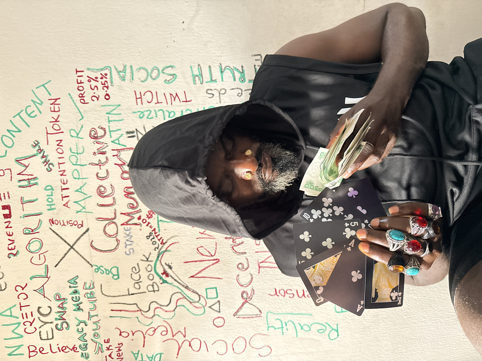 A dark-skinned man is positioned against a light-colored wall, appearing to be resting or reclining, with his head tilted to his right. He wears a black hooded top and black shorts. His eyes are closed or cast downwards, and he has a beard and two small, gold nose studs.
In his left hand, he holds a wad of green banknotes, likely Nigerian Naira, featuring "500" denominations. His right hand holds several black playing cards, fanned out, revealing two Aces of Clubs, a 5 of Clubs, and a 10 of Clubs. The backs of some cards feature an ornate, gold-toned design. Both of his hands are adorned with multiple large, ornate rings set with colorful stones, including red, turquoise, and amber.
The wall behind him is covered with numerous words and symbols in red, green, and black markers, resembling graffiti or a whiteboard brainstorming session. Visible green text includes "CONTENT," "ALGORITHM," "SHARE," "MAPPER," "Collective," "Base," "STAKE," "Book," "Social," "PROFIT 2.5%," "TWITCH," and "TRUTH SOCIAL." Red text includes "REVENUE," "NWA," "CETOR," "SWAP," "LEGACY MEDIA," "YOUTUBE," "Believe," "POSITION," "HOLD," "ATTENTION TOKEN," "Memory," "ATTN=10$," "2028," "New," "Decensor," "Reality," and "PROFIT 0.25%." A large black "X" is drawn in the center, and various geometric shapes and wavy lines are also present. The scene is brightly lit, casting some shadows from the man onto the wall.
