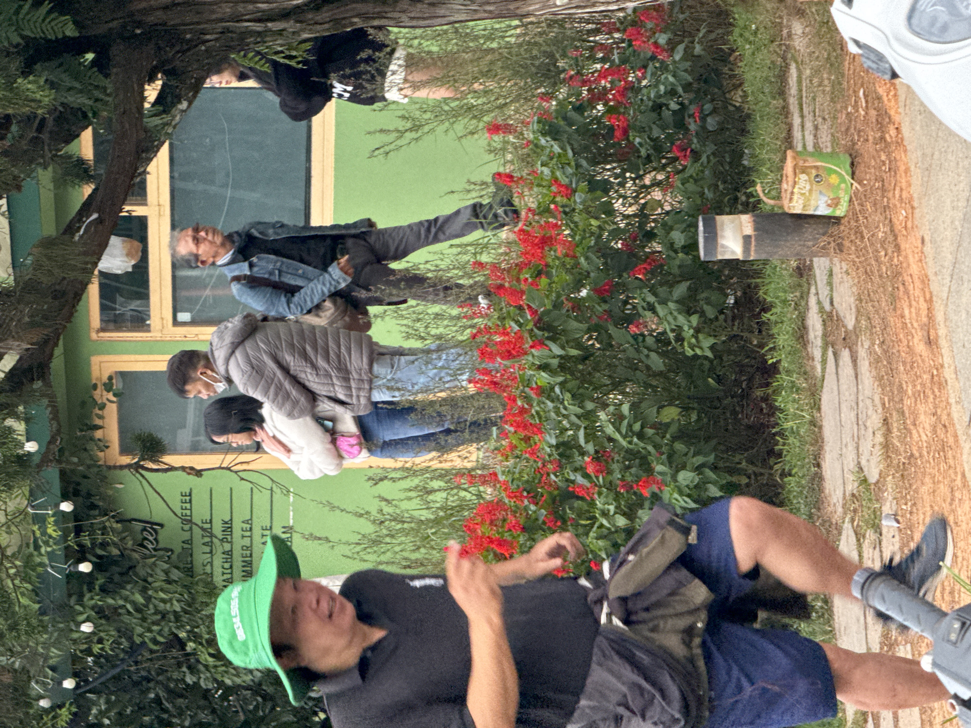 The image depicts a street scene in Da Lat, Vietnam, near a building with a bright green facade and yellow window frames. In the foreground, a person wearing a green bucket hat and a black polo shirt is partially visible, walking to the left. They are wearing blue shorts and a dark jacket tied around their waist. Behind them, a woman in a light-colored sweater and jeans stands with her arms crossed, next to a man in a grey jacket and jeans, who is also wearing a face mask. Further back, another man in a denim jacket and dark pants is visible. A line of red flowers runs along the front of the green building, with tall green foliage behind them. A lamppost and a small green bucket with a handle are situated on the ground in the lower right portion of the image. The overall setting suggests an outdoor cafe or shop area in Da Lat.