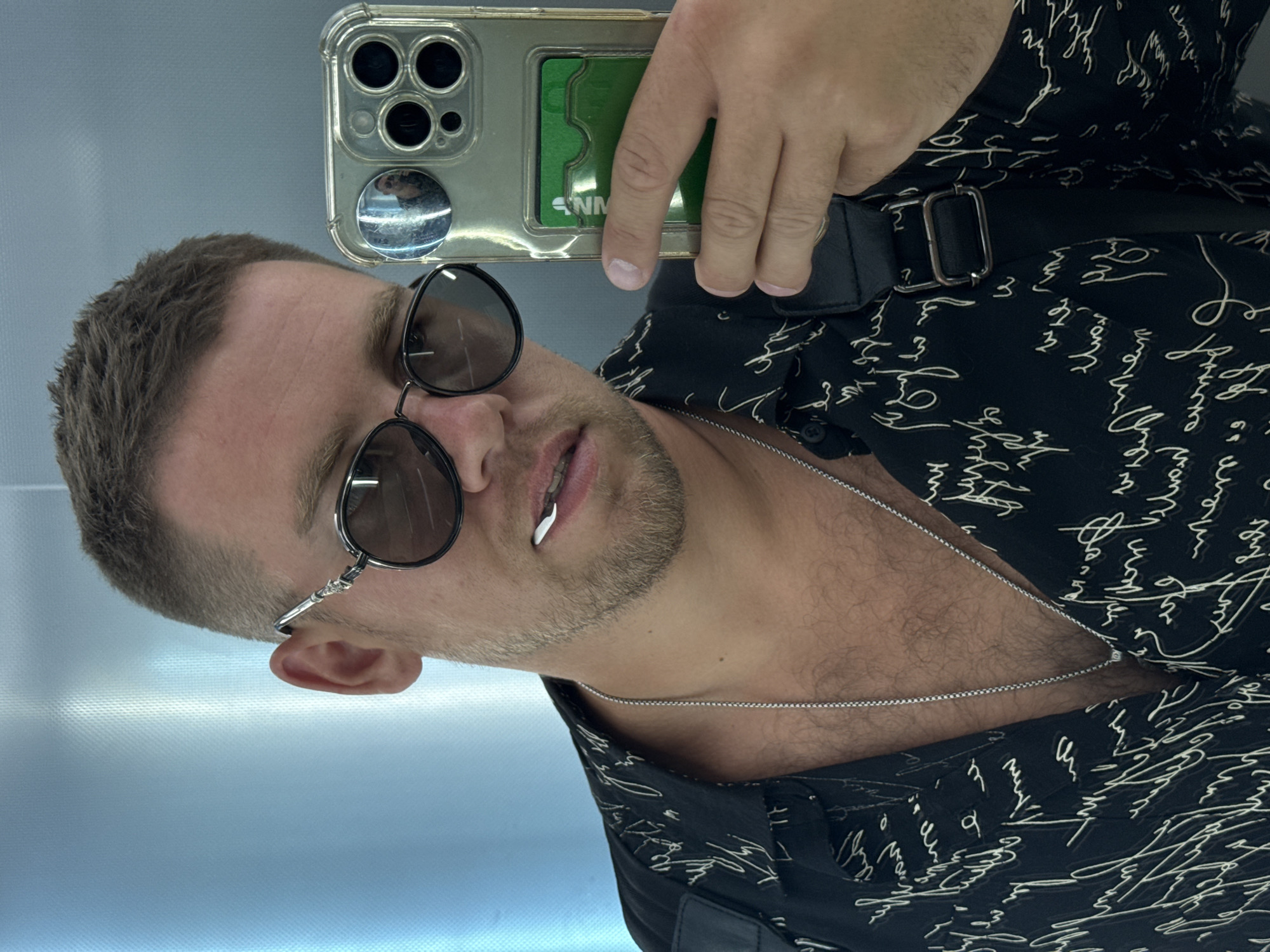 A close-up selfie, captured in a reflective surface, features a man looking towards the camera with his head tilted. He has short, neatly trimmed dark hair and light stubble on his face. He is wearing round sunglasses with dark lenses and thin frames. His mouth is slightly open, revealing a metallic, decorative accessory on his lower lip. A silver chain is visible around his neck, resting on his upper chest.
The man is dressed in a black shirt adorned with an all-over white script-like pattern, which is stylized and appears to be handwritten text, though largely illegible. A black strap with a silver rectangular buckle is visible over his shoulder, suggesting a bag or sling.
He is holding an iPhone, identifiable by its camera array, in a clear case with his right hand, positioned above his head. A green card, with the partially visible text "NM", is placed within the clear phone case. A small, circular mirror attachment on the phone case reflects a blurry image of the man. The background is a uniform light blue-grey reflective surface, indicating an indoor setting. The man's expression is neutral.