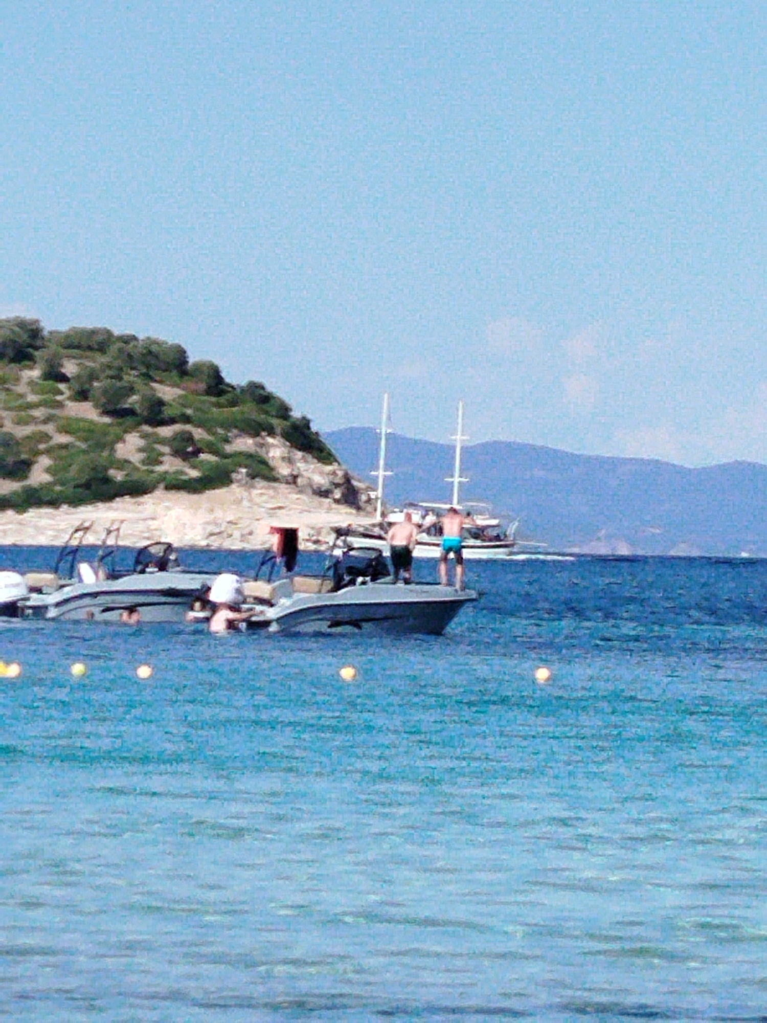 In the foreground, the clear, bright blue water of the Aegean Sea is calm, with subtle ripples reflecting the sunlight. Several yellow buoys are visible floating on the surface, spaced apart.
In the midground, two motorboats are moored in the water. The boat on the left is partially obscured by the boat on the right. The boat on the right features several people. One individual, wearing blue shorts, stands on the edge of the boat, facing away from the camera. Another person, wearing dark shorts, is also on the boat, further back. A third person in white is leaning over the side of the boat. The boats appear to be recreational or speedboats.
In the background, a larger, white sailboat with two masts is visible on the horizon, moving to the right. Beyond the water and boats, a hilly coastline is depicted. The hills are covered in dense green vegetation, with some exposed rocky areas. Further in the distance, hazy blue mountains form the backdrop against a clear, light blue sky. The overall scene suggests a tranquil day at sea, likely in a coastal area of Greece. The specific location is within the Sithonia Municipal Unit, known for its beautiful beaches and clear waters.